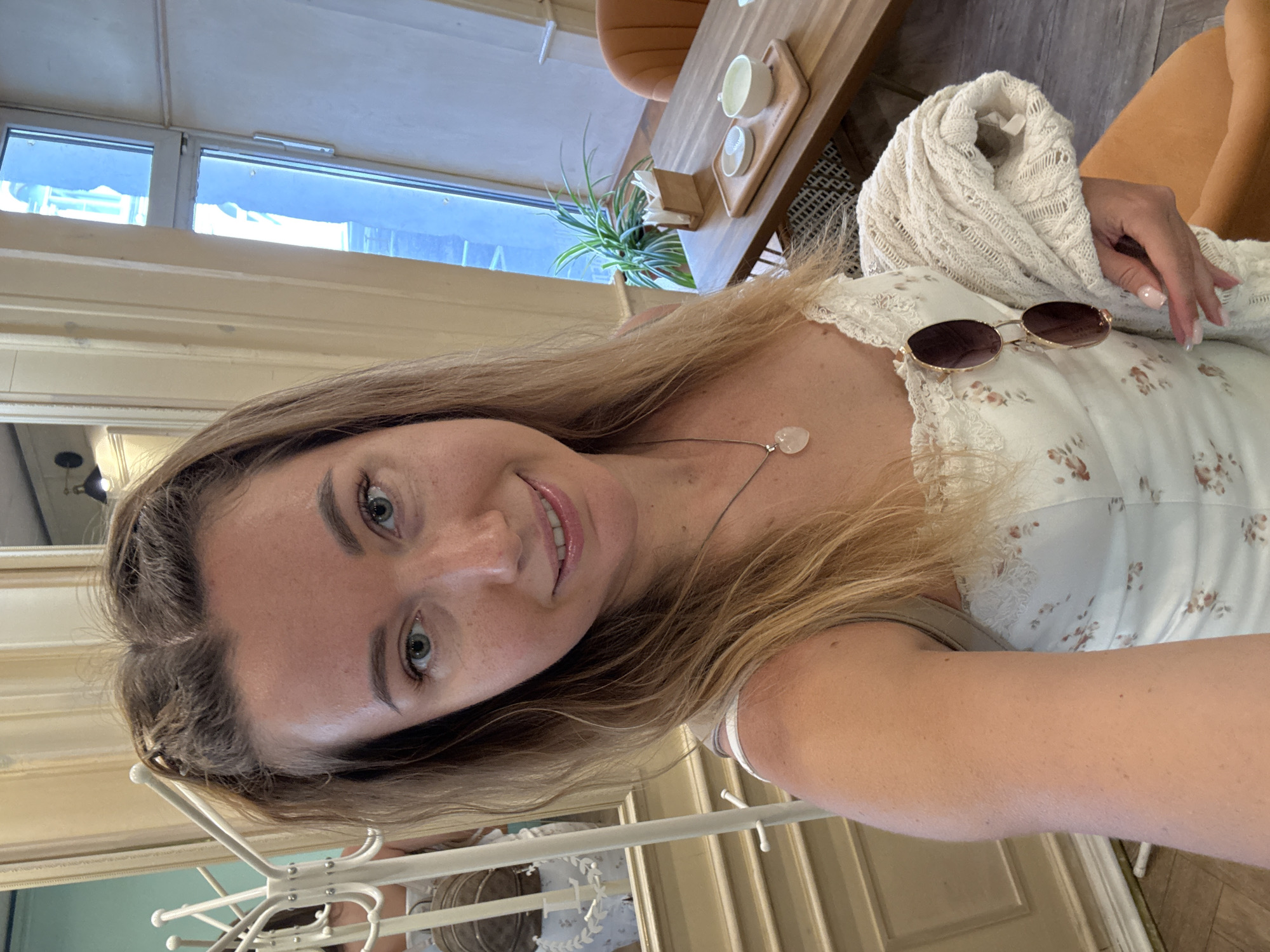 A fair-skinned woman, approximately 25-35 years old, with long blonde hair and light blue-green eyes, is centered in a selfie-style portrait. She has a slight smile, revealing closed lips, and visible freckles across her nose and cheeks. Her hair is parted slightly off-center and appears somewhat disheveled at the roots. She is wearing a white sleeveless dress with a floral pattern and lace trim, featuring thin straps. A silver-colored necklace with a light pink, heart-shaped pendant is visible around her neck. Brown-tinted sunglasses are hooked onto the front of her dress. Her left hand, with light pink fingernail polish, holds a light cream-colored, cable-knit sweater or cardigan draped over her arm.
The background shows an indoor setting, likely a cafe or restaurant. To the left, a white, ornate coat rack or hat stand is positioned, and behind it, a large mirror reflects another person, partially obscured, wearing a light-colored outfit and a small brown backpack. To the right of the woman, a wooden table is visible with a light-colored tray holding two small white dishes or cups. A wooden napkin dispenser and a potted green plant are also on the table. An orange upholstered chair is partially visible behind the table. Further to the right, a large window reveals a bright outdoor scene with faint building outlines. The floor has a subtle geometric pattern, and the walls feature light-colored paneling.