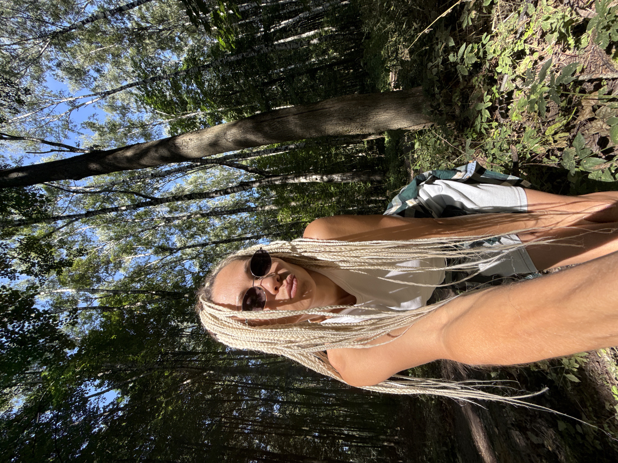 A woman with blonde braids and sunglasses is taking a selfie in a forest. She is wearing a white tank top and green and blue plaid shirt. She is looking at the camera with her lips pursed. The forest has tall trees and sunlight filtering through the leaves. The ground is covered in leaves and undergrowth. The image appears to be taken during the daytime. The location is Dumino, Russia.