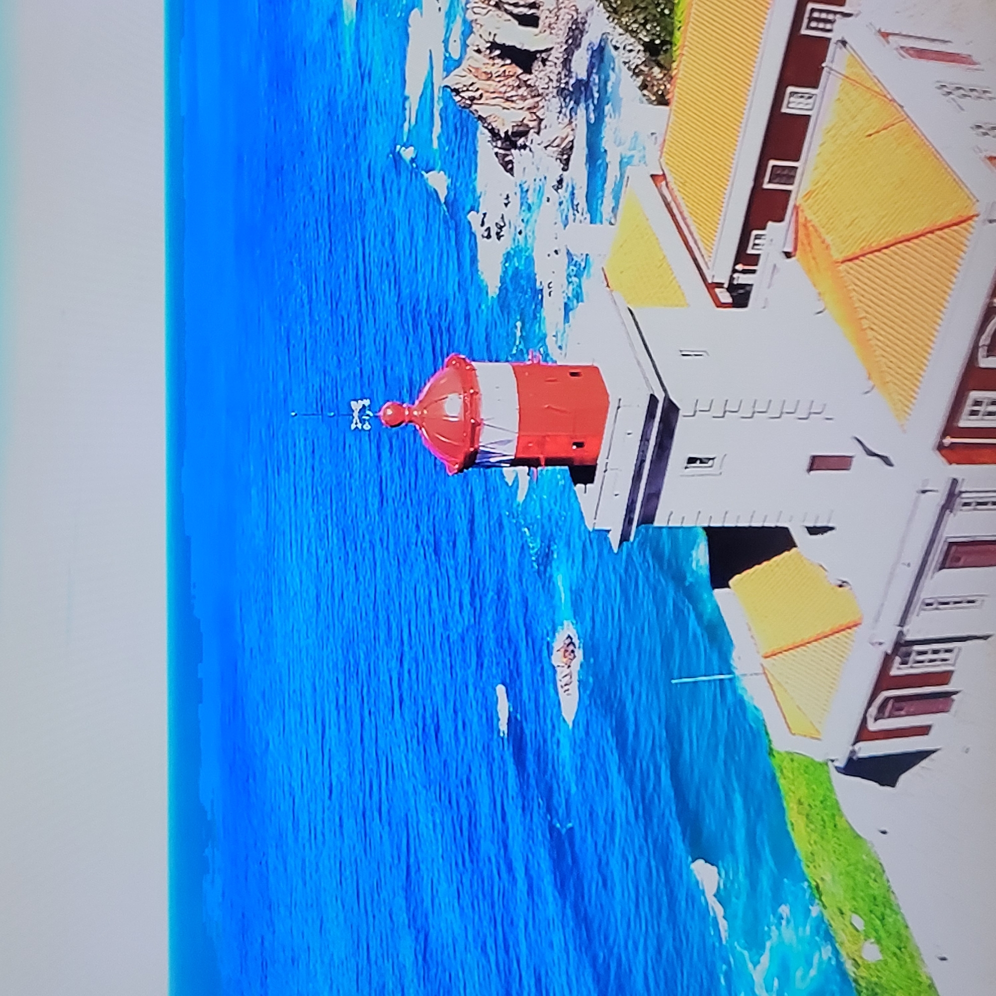The image shows a coastal scene with a lighthouse. The lighthouse has a red top and a white base. The buildings attached to the lighthouse have yellow roofs. The scene includes the ocean, rocky cliffs, and some greenery. There are also boats visible in the water. The image does not appear to be related to Yerevan, Armenia.