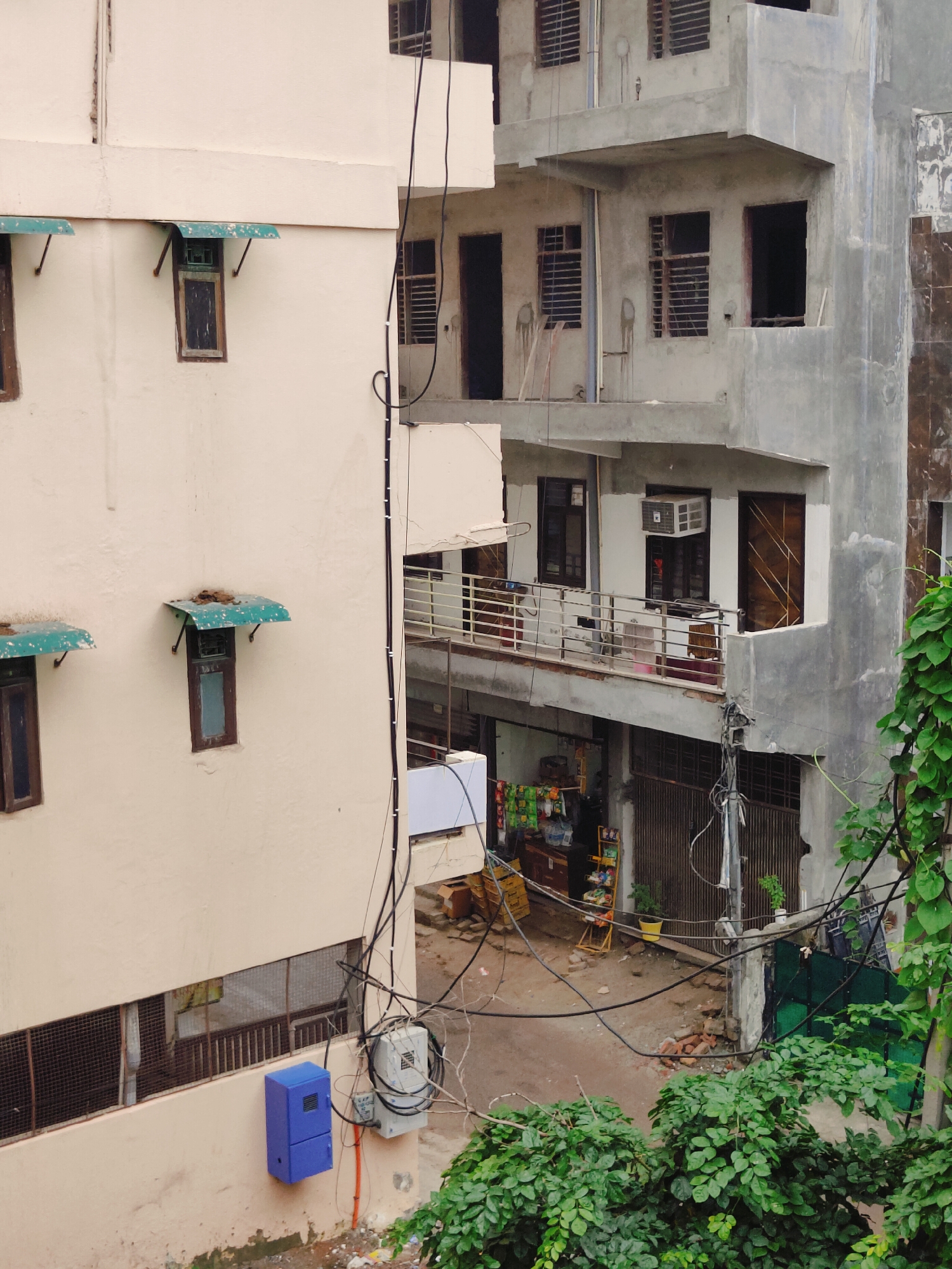 The image shows an urban scene in Gurgaon, India, featuring multi-story buildings under construction or renovation. On the left, a cream-colored building has several windows with green awnings. The building on the right is a concrete structure with exposed pipes and unfinished concrete surfaces, displaying various windows, some with metal grates, and balconies with railings. An air conditioning unit is visible on one of the balconies. Below the balconies, there is a narrow street or alleyway with a small shop displaying various goods. Electrical wires are strung across the scene, connecting to electrical boxes mounted on the buildings. Lush green foliage is present in the foreground and on the right side of the image, partially obscuring the lower parts of the buildings. The ground is a mix of dirt and scattered bricks.