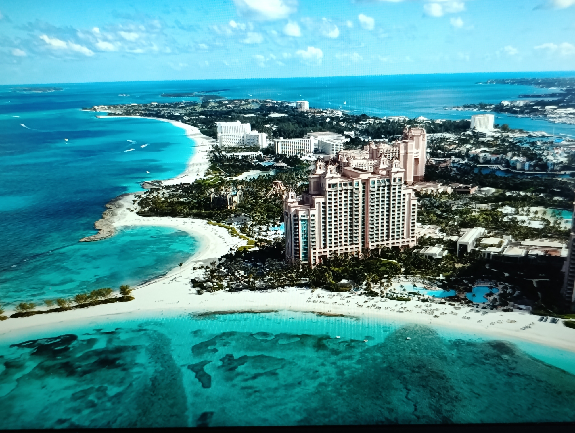This is an aerial, wide-angle view of a tropical resort area. The scene is dominated by vibrant turquoise and blue ocean waters meeting white sandy beaches and lush green tropical vegetation. Several large, multi-story resort buildings with light-colored facades are visible. One prominent pink tower building with many windows and distinct architectural features stands out. In the foreground, the shallow ocean floor is visible through the clear water, showing reefs and rock formations. Boats are scattered across the water. The sky is blue with scattered white clouds. The location is not in Dubăsari town council, Moldova, as that is a landlocked area in Europe, and this image clearly depicts a tropical coastal resort.