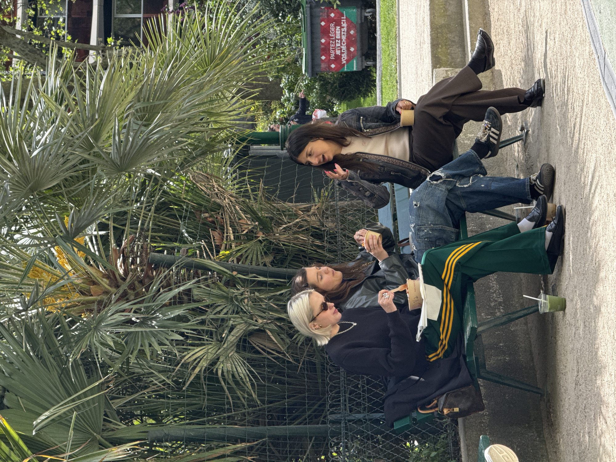 Three young women are casually seated on a green metal bench in an outdoor park setting in Paris, France, on a bright, sunny day. The scene is dominated by lush, palm-like foliage and a dark green fence.
The woman at the top, with dark hair, wears a dark denim jacket over a light top and dark wide-leg trousers, paired with black loafers and sneakers. She is smiling broadly and gesturing with her hand, holding a paper cup, appearing engaged in lively conversation. The middle woman, also with dark hair, is dressed in a black leather jacket and holds a small container. The woman at the bottom, with blonde hair and sunglasses, wears a black long-sleeved top, dark green track pants with yellow stripes, and black shoes. A silver chain necklace adorns her neck, and a Louis Vuitton Speedy bag rests next to her on the bench. She is holding a container or paper bag with a fork. A green cup with a straw is on the ground next to the bench.
The women appear relaxed and are interacting, suggesting a social gathering. In the background, a green public trash bin is visible, bearing the French text: "PARTEZ LÉGER, JETEZ BIEN VOS DÉCHETS ICI!" (TRAVEL LIGHT, THROW YOUR WASTE HERE!). The bright sunlight casts clear shadows, indicating a pleasant, clear day.