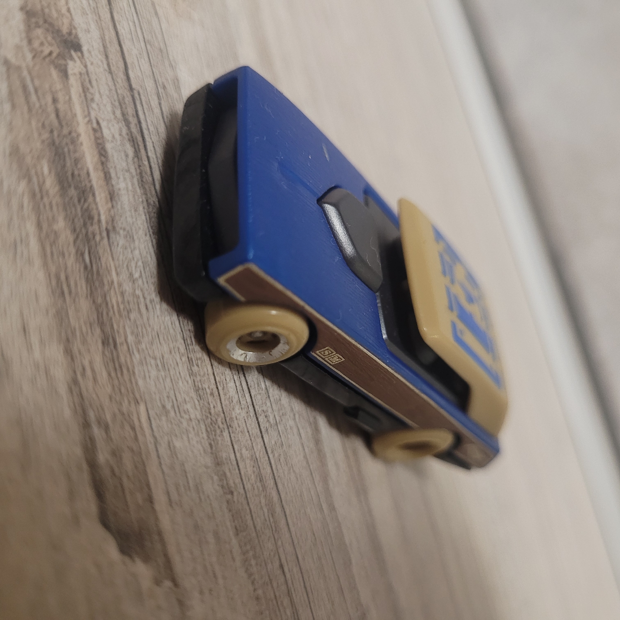 A toy car is placed on a wooden surface. The car is predominantly blue with a tan roof and brown detailing on the sides. The tan roof has blue lettering. The wheels are tan with silver rims. The surface is a light-colored wood with visible grain. The background is out of focus, showing a lighter colored surface at an angle. The image is a close-up shot of the toy car, angled from the front right.