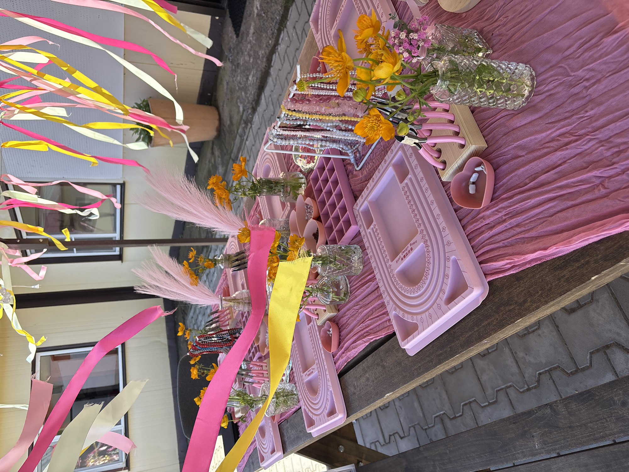 The image depicts an outdoor table set up for a jewelry-making activity, possibly a children's party. The table is covered with a crinkled pink cloth. On the table are several pink jewelry design boards, bead trays, and small containers of beads. There are also vases of orange and pink flowers, as well as pink feather decorations. Ribbons in pink, yellow, and white are strung overhead, adding a festive atmosphere. In the background, a building with windows is visible, and the ground is paved with interlocking stones. The overall scene suggests a creative and colorful event. The location is not specified in the image or the provided crops, therefore it cannot be identified as Birobidzhan, Russia.