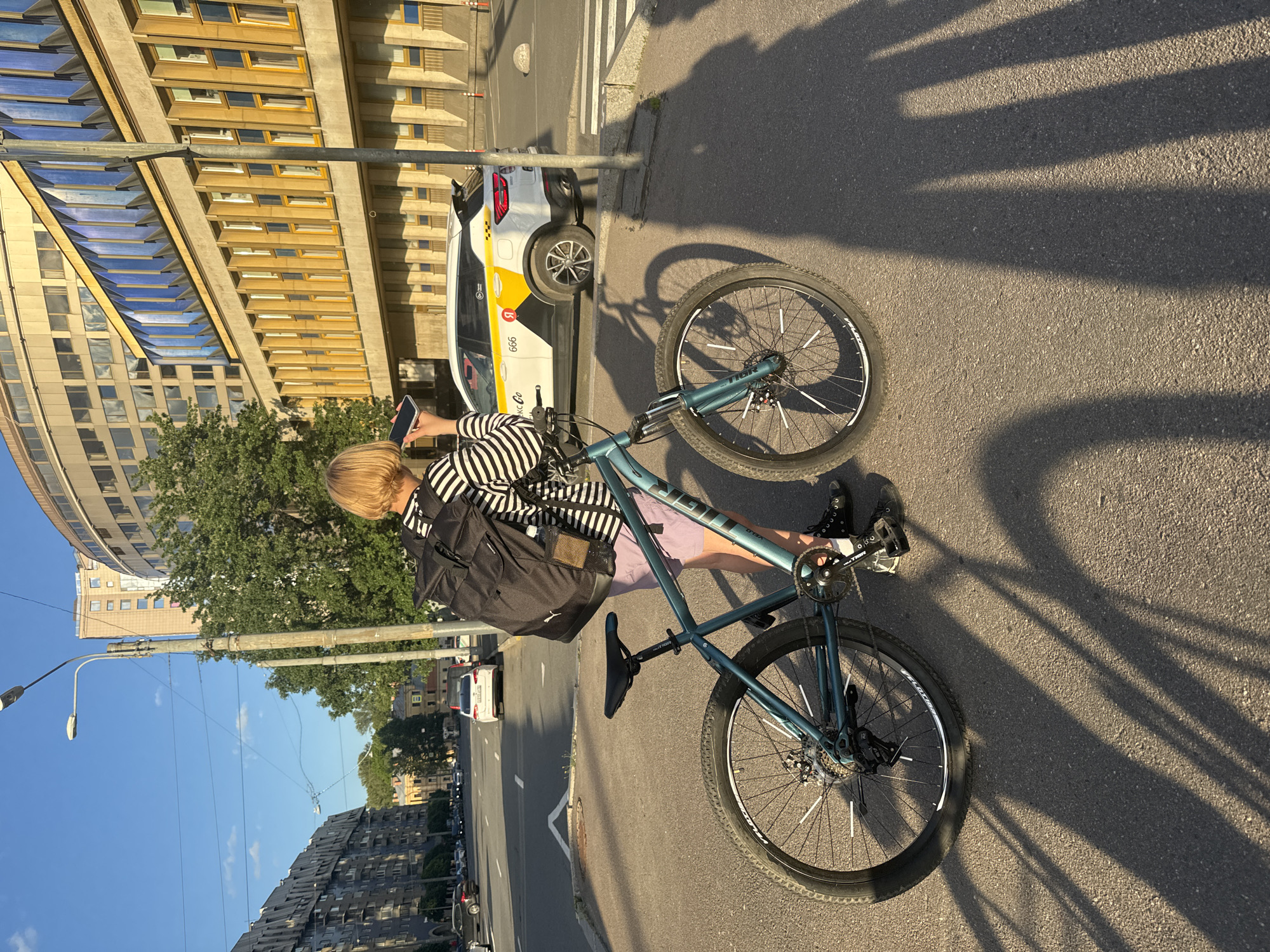 A person with blond hair, wearing a striped shirt and shorts, stands next to a teal bicycle on a paved street in Saint Petersburg, Russia. The person is holding a mobile phone to their ear, as if on a call. The bicycle has a black backpack attached to its frame. In the background, a white taxi with yellow and black markings is parked on the street, and several buildings line the road. The scene is illuminated by the warm glow of the setting sun, casting long shadows across the pavement.