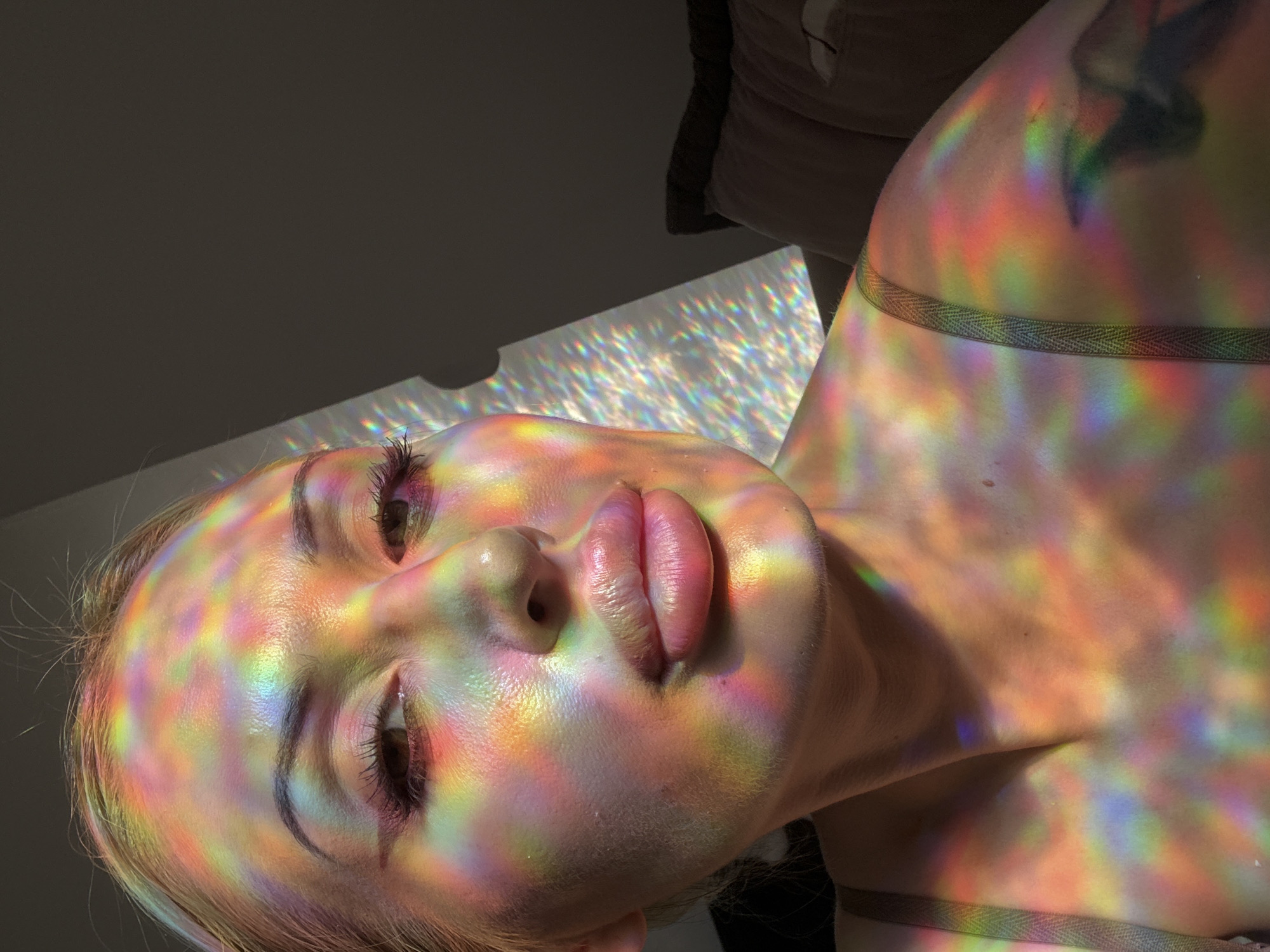 A close-up, medium shot captures a woman's face and upper body bathed in prismatic light. The light, casting colorful reflections across her skin, creates a rainbow effect on her forehead, cheeks, nose, lips, neck, and arm. Her eyes are partially closed, and her lips are slightly parted. She has makeup on, including pink eyeliner. A tattoo is visible on her arm, obscured by the light. The background is dark and indistinct, with what appears to be a pillow and a dark wall. The scene is lit by natural light filtering through a prism or reflective surface. The location is unknown.