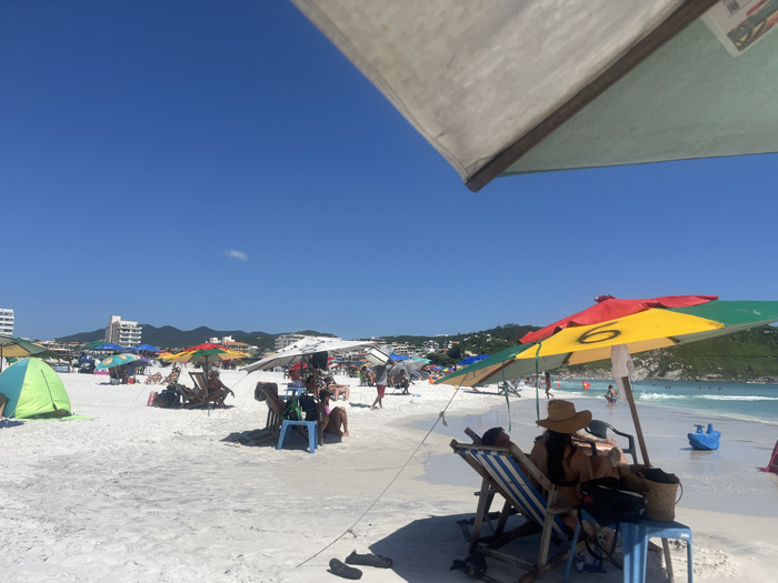 The image displays a vibrant and bustling beach scene on a clear, sunny day. The perspective is taken from under a light-colored beach umbrella, a corner of which is visible in the upper right portion of the frame.
In the foreground, slightly to the right, two beach chairs are set up. One chair is occupied by a person seen from behind, wearing a straw hat and appearing to relax under the shade of a brightly colored umbrella. This umbrella features distinct sections of red, yellow, and green fabric, with the number "6" clearly visible in black paint on its yellow section. Next to these chairs, a blue plastic stool holds a woven bag, and a pair of black flip-flops lie on the pristine white sand. To the left, a light green and blue tent is pitched on the sand.
The expansive midground reveals a wide stretch of brilliant white sand, dotted with numerous colorful beach umbrellas and smaller tents, indicating a popular and well-attended beach. Many people are scattered across the sand, relaxing in chairs, walking, or standing. In the background, beyond the immediate beach area, multi-story white buildings are visible, suggesting a developed coastal town or resort. These buildings are backed by rolling hills covered in lush green vegetation.
The clear, calm ocean waters, a beautiful turquoise blue, stretch across the horizon. Several individuals can be seen in the water, swimming or wading close to the shore. A small, bright blue object, possibly a child's toy or a small boat, floats near the water's edge. The sky above is a brilliant, cloudless blue, save for a single small, wispy white cloud floating high up, reinforcing the impression of perfect beach weather. The overall scene conveys a lively atmosphere of leisure and enjoyment.