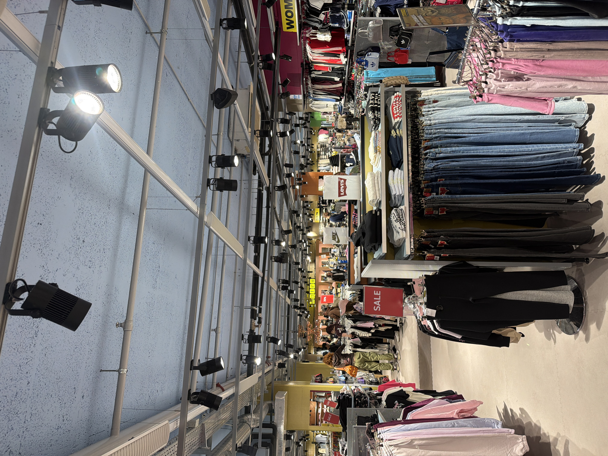 The image depicts the interior of a clothing store in Spreitenbach, Switzerland. A grid of metal beams and track lighting fixtures covers the ceiling. Rows of clothing racks display various items, including jeans, sweaters, and athletic wear. A "SALE" sign is visible among the clothing. Shoppers are present, although details are obscured.