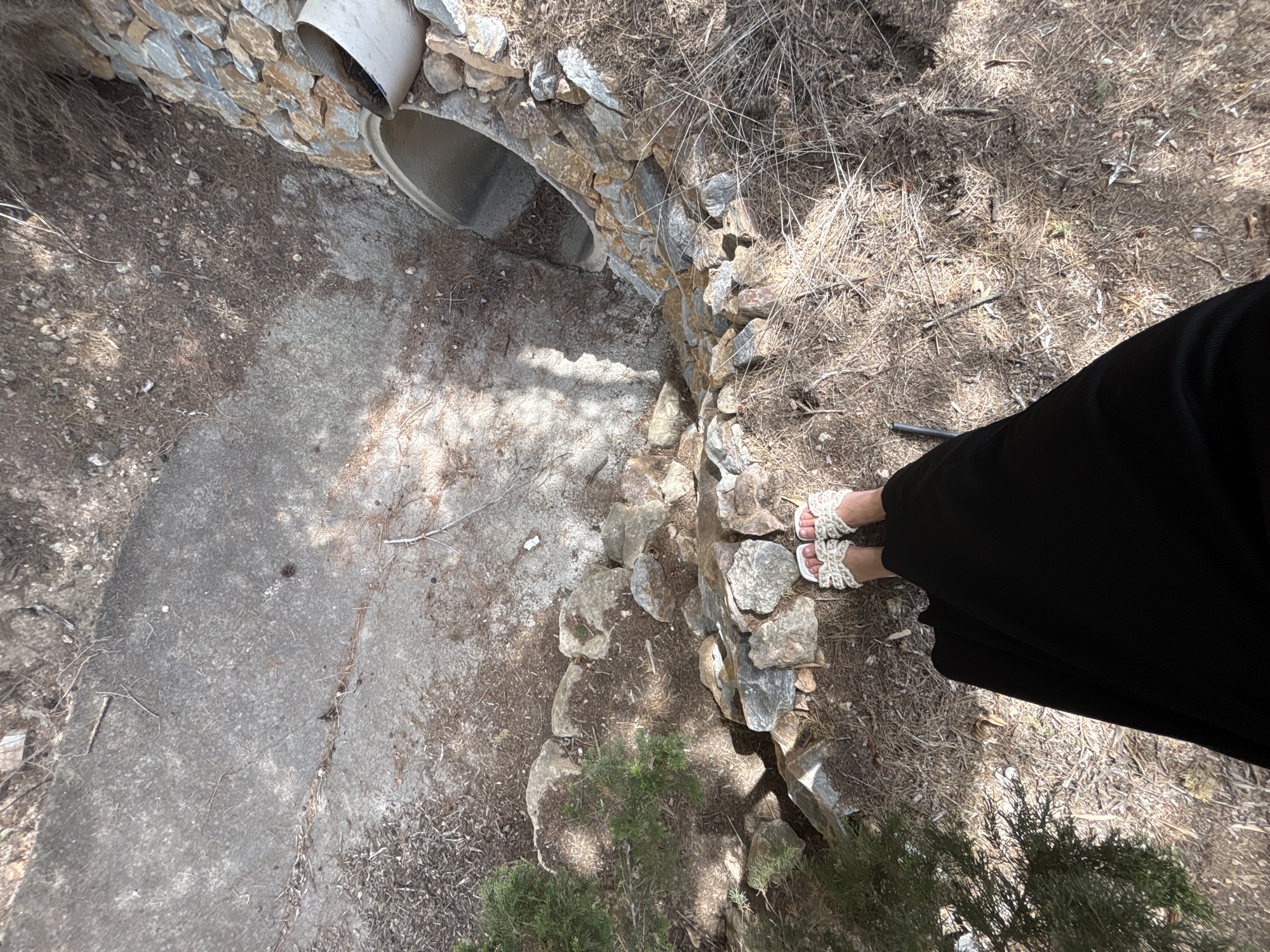 The media file shows a high-angle view of an outdoor scene, likely in Orihuela, Spain. The foreground features the lower half of a person wearing a long black skirt and white, crocheted sandals with a beaded detail. The person is standing on dry, brown grass and dirt, interspersed with small rocks and twigs.
The scene also includes a concrete path or surface to the left, which appears worn and somewhat cracked. Adjacent to the path is a stone retaining wall built around a large, concrete culvert pipe that enters the ground at an angle. The culvert is partially obscured by shadows and dry vegetation. The sun appears to be high in the sky, suggesting it is midday, and the lighting indicates a clear, dry day. The overall environment appears somewhat arid and rustic. There is no visible text in the image. The focus is on the textures of the ground, the stone work, and the person's attire.