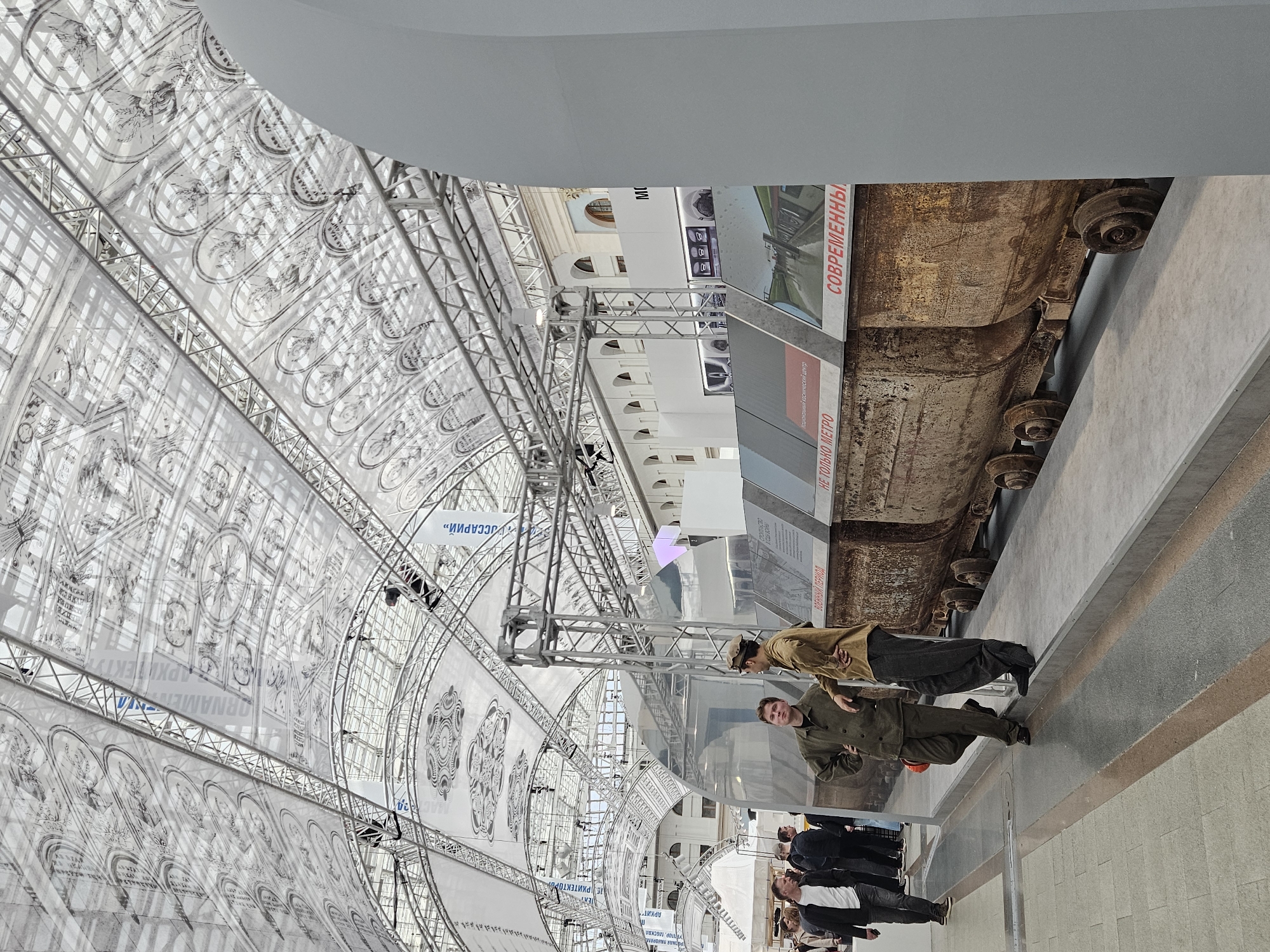 The image captures a scene within the Moscow Metro in Russia, specifically at the Partizanskaya station, which is part of the Moscow Metro's Arbatsko-Pokrovskaya Line. The shot is taken at a low angle, emphasizing the station's architectural details, including a large ceiling adorned with historical and patriotic motifs.
Several people are present in the scene. Two men in green and brown jackets seem to be standing and conversing, while a group of individuals is gathered in the background, some walking and some appearing to be stationary. The lighting in the metro station is bright, showcasing the white and gray color palette that dominates the architectural design.
An old rusty metro train car is also on display. Some signs in Russian are visible.