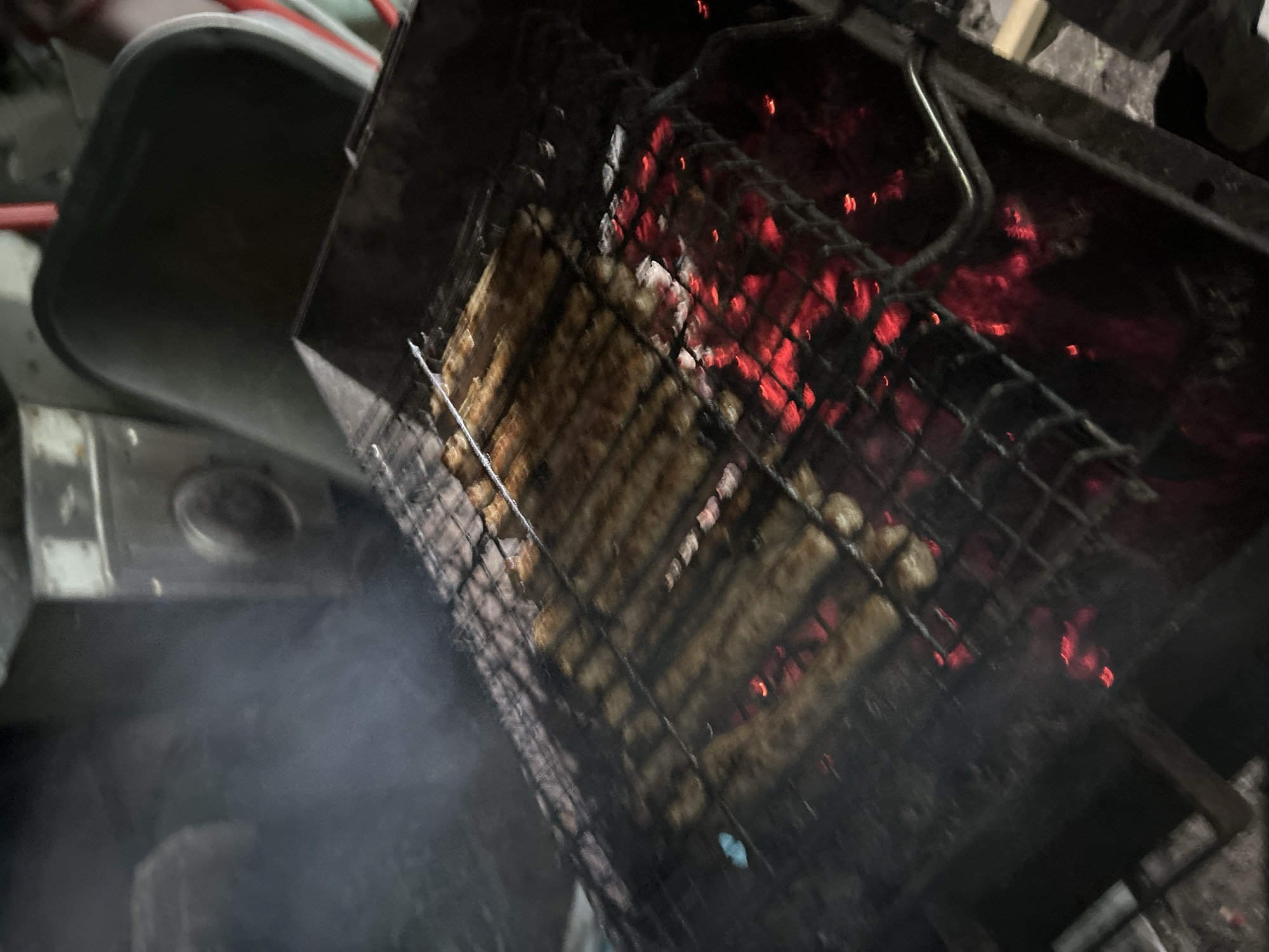 A close-up, overhead, blurry shot shows food being grilled over hot coals. The food, which appears to be sausage links, is laid out on a metal grill grate. Red-hot coals glow beneath the grate, creating a warm light. Wisps of smoke rise from the grill, obscuring parts of the background. The overall scene is dark and appears to be outdoors, possibly in a backyard or campsite. There are no people visible in the image.