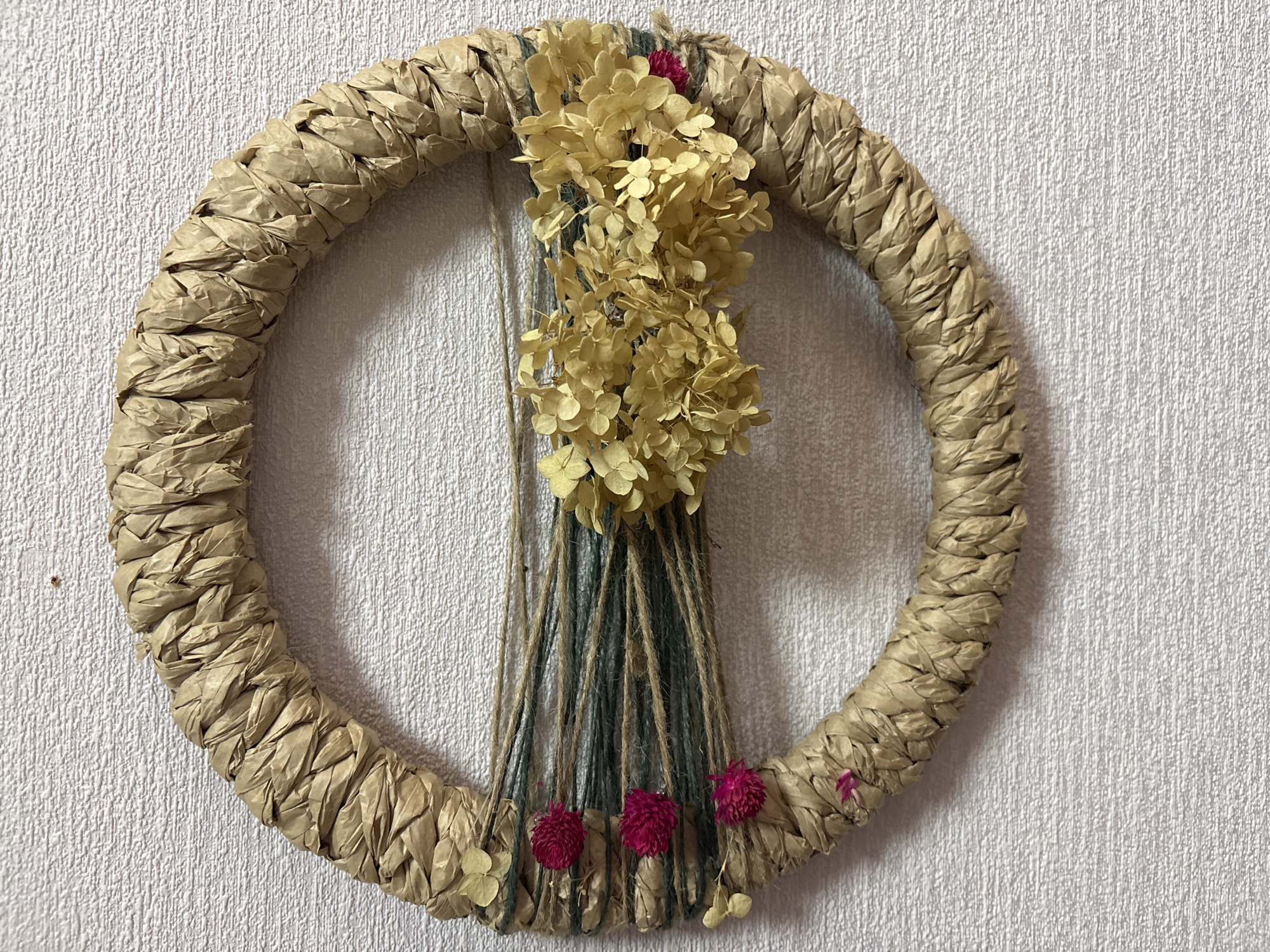 This is a close-up, eye-level, medium shot of a circular wreath made of braided brown paper or straw, hanging on a white textured wall. The wreath is adorned with several bundles of dried yellow flowers and small pink flowers, tied together with twine and green string. The lighting is natural and even, casting subtle shadows on the wall. The overall impression is of a rustic, handmade decoration. The location is identified as Ветрынскі сельскі Савет, Belarus.