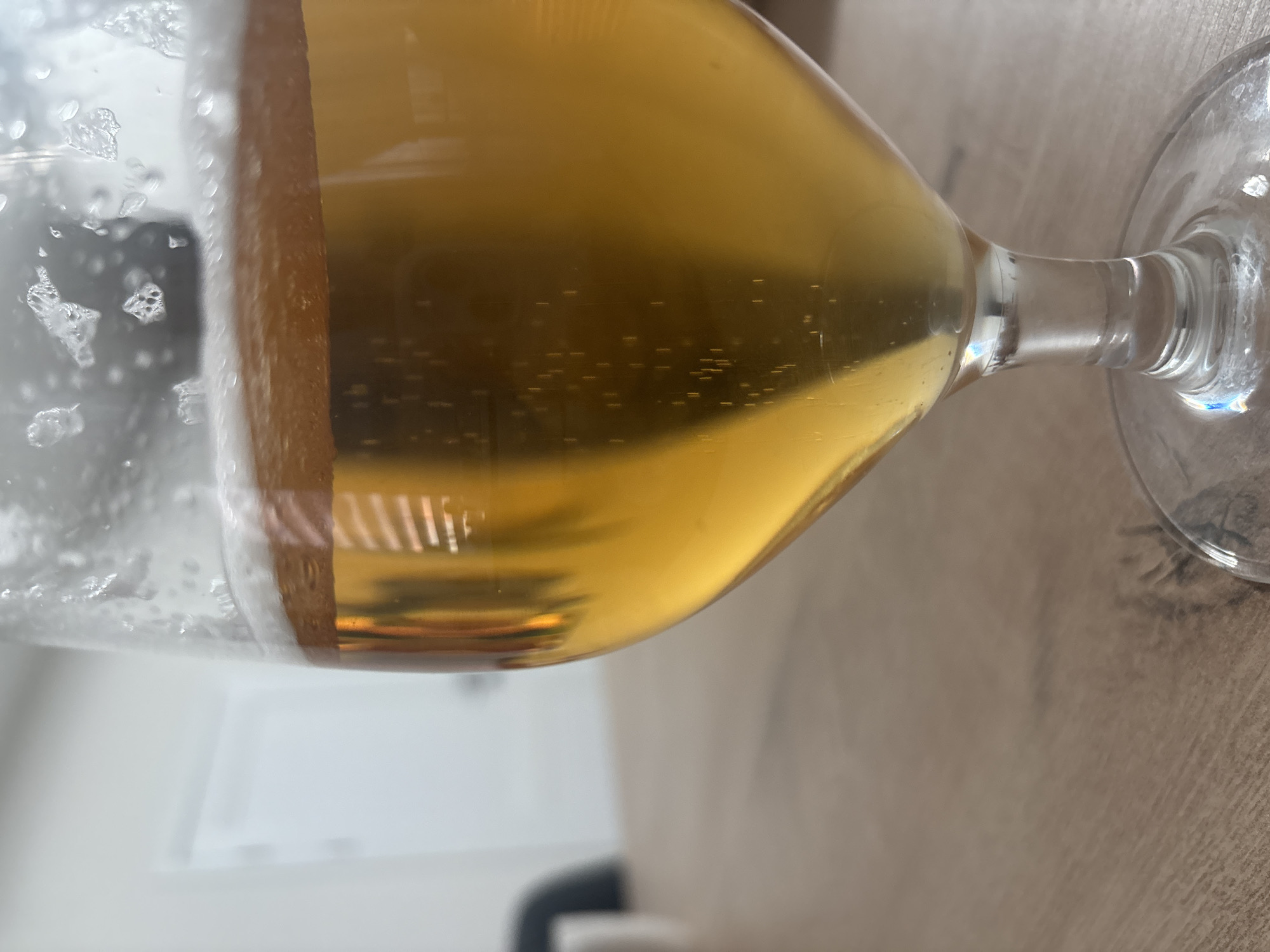 The image is a close-up, eye-level, slightly angled shot of a glass filled with a golden-hued beverage, likely beer, with a foamy head. The glass is positioned on a light brown wooden surface, possibly a table. The background is out of focus, showing a white wall and a doorway or opening. Subtle reflections are visible on the surface of the liquid and the glass. The lighting appears to be ambient, coming from the left and slightly above. The scene does not contain any people or specific recognizable landmarks of Chaikovsky, Russia.