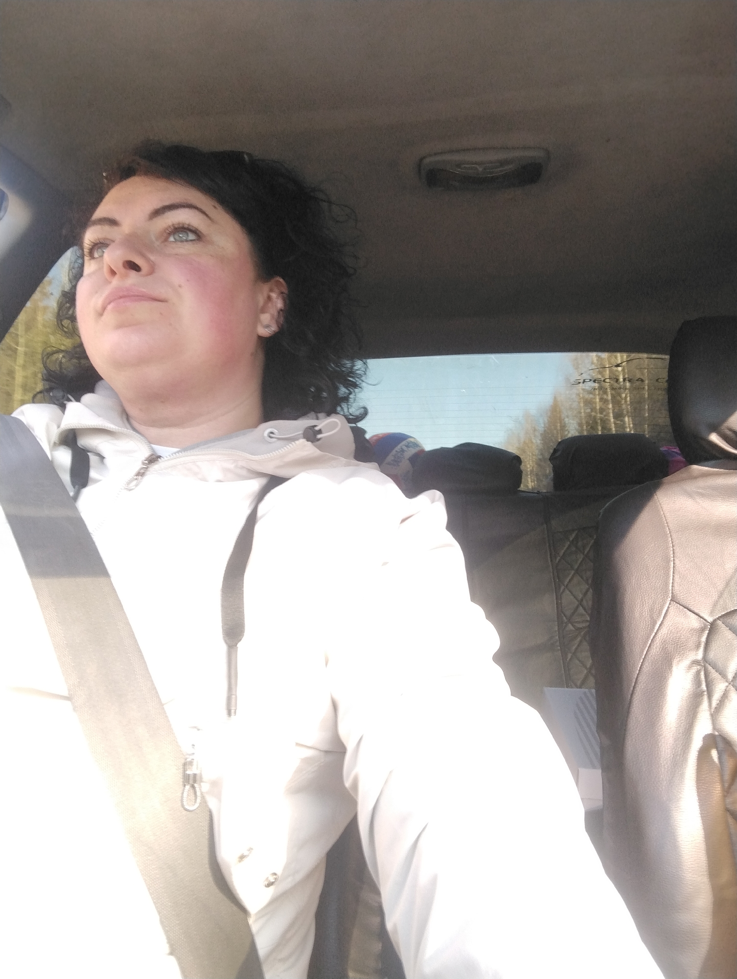 The image features a woman seated in the front of a car, likely in Dvigatel', Russia, under bright daytime conditions. She is positioned towards the left side of the frame, wearing a seatbelt across her chest and a light-colored, possibly white or cream, hooded jacket with a zipper and drawstrings. Her dark, curly hair frames her face, which is turned upwards and slightly to her left, looking out of the car. Her eyes are light-colored, and her cheeks show some redness. A small metallic object, possibly a keyring or carabiner, hangs from the seatbelt strap.
The car's interior is visible, including the light-colored headliner, an overhead light fixture, and dark-colored rear seats with a quilted pattern on the backrest. Through the car's rear window, a clear blue sky and a sparse line of trees, indicative of a forest or wooded area, are visible in the distance. This suggests the car is on a road adjacent to a natural environment. Bright sunlight illuminates the scene, causing some overexposure on the right side of the woman's jacket and the car's interior.
On the upper right corner of the rear window, faint white text reads "SPECTRA C", which appears to be part of a car model name or a sticker. The overall activity depicted is a person traveling or pausing in a car during a sunny day, with the woman appearing to observe something outside or above. The clear weather and strong sunlight are notable details.