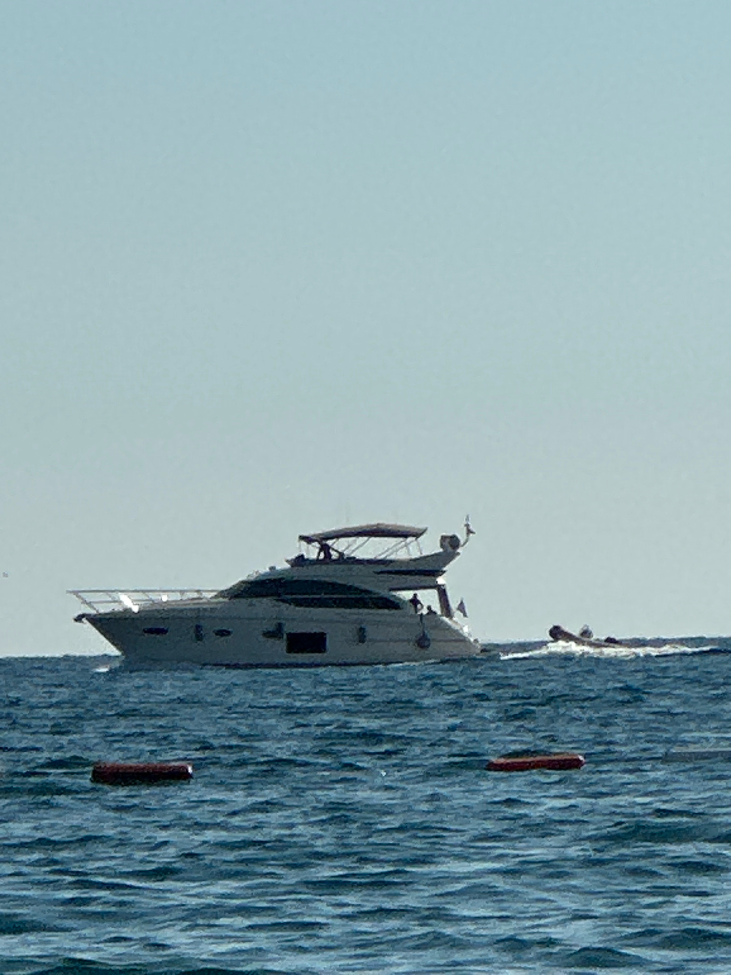 A large white yacht with a dark hull is moving through moderately choppy blue water under a clear, pale blue sky. The yacht has multiple levels and a dark canopy over the upper deck. A small inflatable boat or tender is visible behind the yacht, creating a wake. Two red cylindrical buoys are floating in the water in the foreground. The scene is taking place at sea, not within the city of Boreti, Montenegro.
