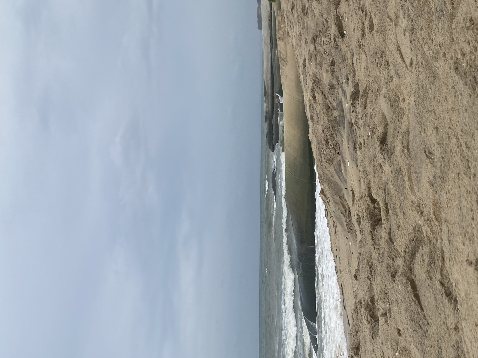 This is a landscape photograph of a beach in Mũi Né, Vietnam, on a cloudy day. The foreground is dominated by the sand of the beach, with footprints and ripples from the wind. In the middle ground, large, dark, smooth rock formations emerge from the sand and into the ocean. The ocean is rough, with white waves crashing against the rocks and shoreline. A person in a white shirt is visible standing on one of the rock formations, facing the ocean with their arms raised. In the background, the horizon is indistinct due to the overcast sky, with a faint outline of land or buildings on the far right. The overall atmosphere is somewhat subdued and moody, consistent with the cloudy weather.