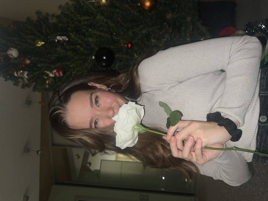 The image depicts a young female indoors in Krasnoyarsk, Russia. She has light brown hair and wears a light grey ribbed long-sleeved top and dark pants. She holds a single white rose with a green stem to her nose, smiling and looking directly at the camera. A black scrunchie is visible on her left wrist, and she wears gold hoop earrings. To her right, a green artificial Christmas tree stands, decorated with various ornaments including red, gold, and black baubles, small Santa figures, and stockings. The environment is an interior space illuminated by warm artificial light.