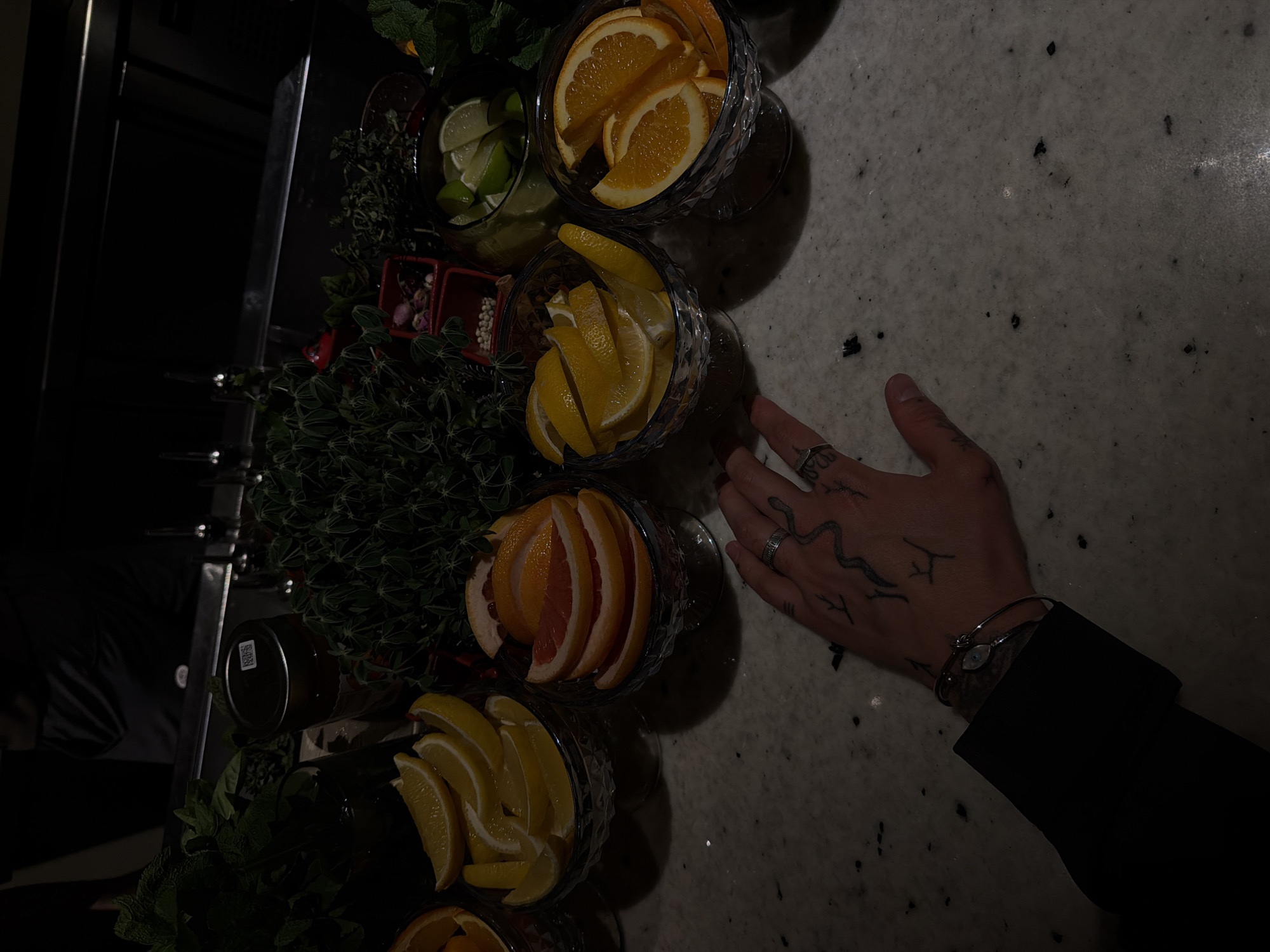 The image shows a collection of glass containers filled with citrus fruits such as lemons, limes, oranges, and grapefruit, and herbs like mint. A person's hand is visible with tattoos and rings, and part of a black sleeve. The location is unclear, Yerevan is a suggestion.