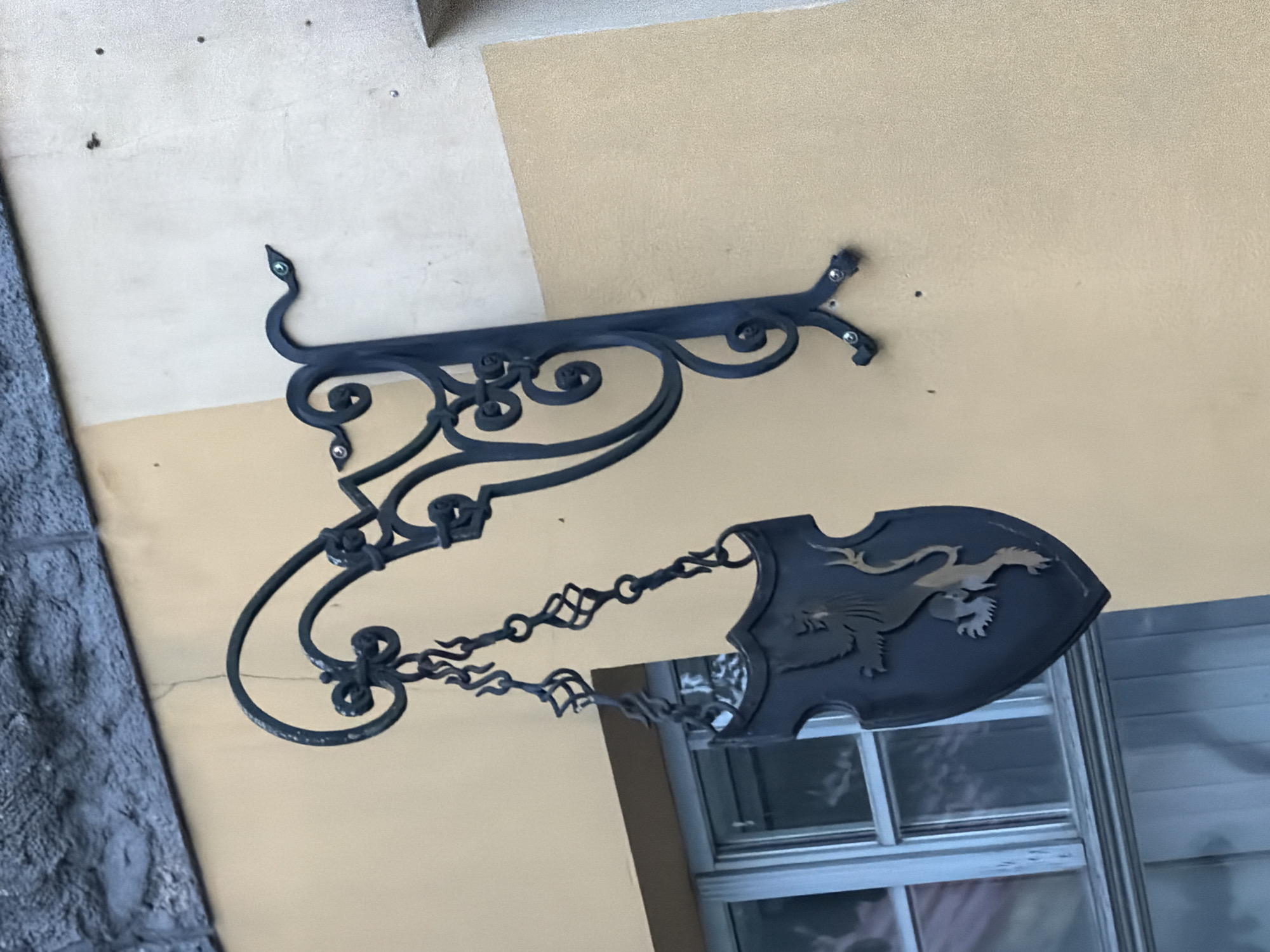 An ornate, dark metal sign hangs from a building's exterior wall in Ventspils, Latvia. The sign features decorative scrollwork and a shield-shaped emblem with a lion depicted on it. The building wall is painted a light yellow color, and a textured gray stone structure is visible above the wall. A window with a light-colored frame is situated to the left of the sign. The scene appears to be an outdoor setting in an urban environment.