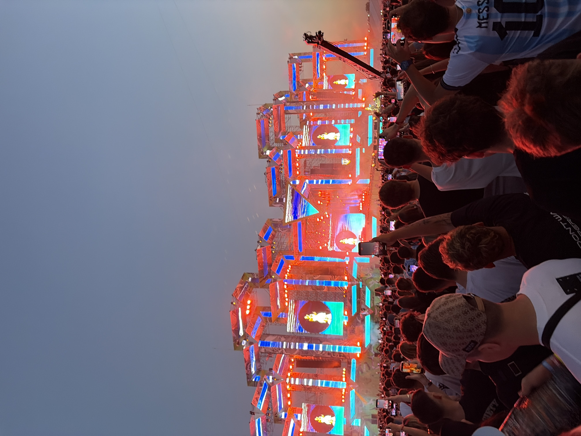The image depicts a large outdoor music festival at sunset in Costinești, Romania. A massive, elaborately constructed stage with numerous screens and lighting fixtures dominates the background, illuminated by vibrant red and blue lights. The stage displays images of a performer. In the foreground, a dense crowd of thousands of people is visible, many holding up their mobile phones to record or photograph the event. The attendees are predominantly young and dressed in casual attire. The atmosphere appears energetic and festive, typical of a summer music festival. The sky is a clear, pale blue, transitioning to a warmer hue near the horizon, indicating the time of day.