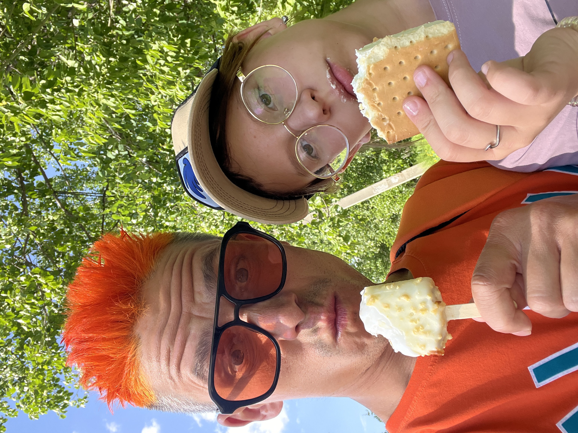 Two people are depicted in a photo taken outdoors. The person on the left has orange hair, wears black framed sunglasses, and an orange t-shirt. The person holds a partially eaten ice cream popsicle. The person on the right wears gold-rimmed glasses and a baseball cap, and is eating a cracker with white icing. The background includes green leaves and a blue sky. The exact location in Chaikovsky, Russia cannot be determined from the image.