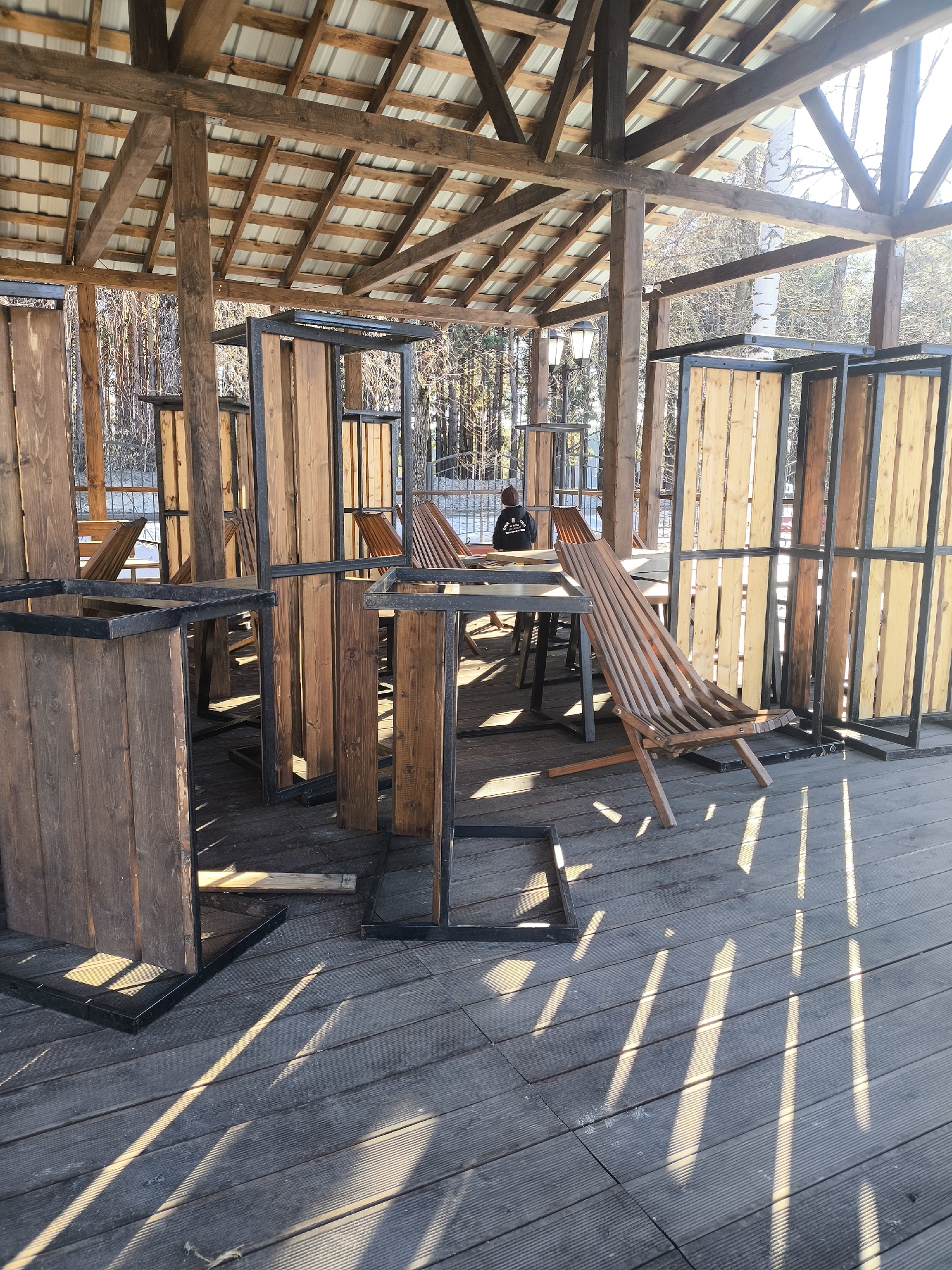 The image shows an outdoor seating area, likely a restaurant or cafe, in Chaikovsky, Russia. The seating area is covered by a wooden structure with a metal roof. Inside the area are wooden tables and chairs, along with wooden partitions with metal frames, possibly for privacy. A person is seated at one of the tables. Sunlight streams through the openings in the structure, creating strong shadows on the wooden deck floor. The trees are visible through the partitions.