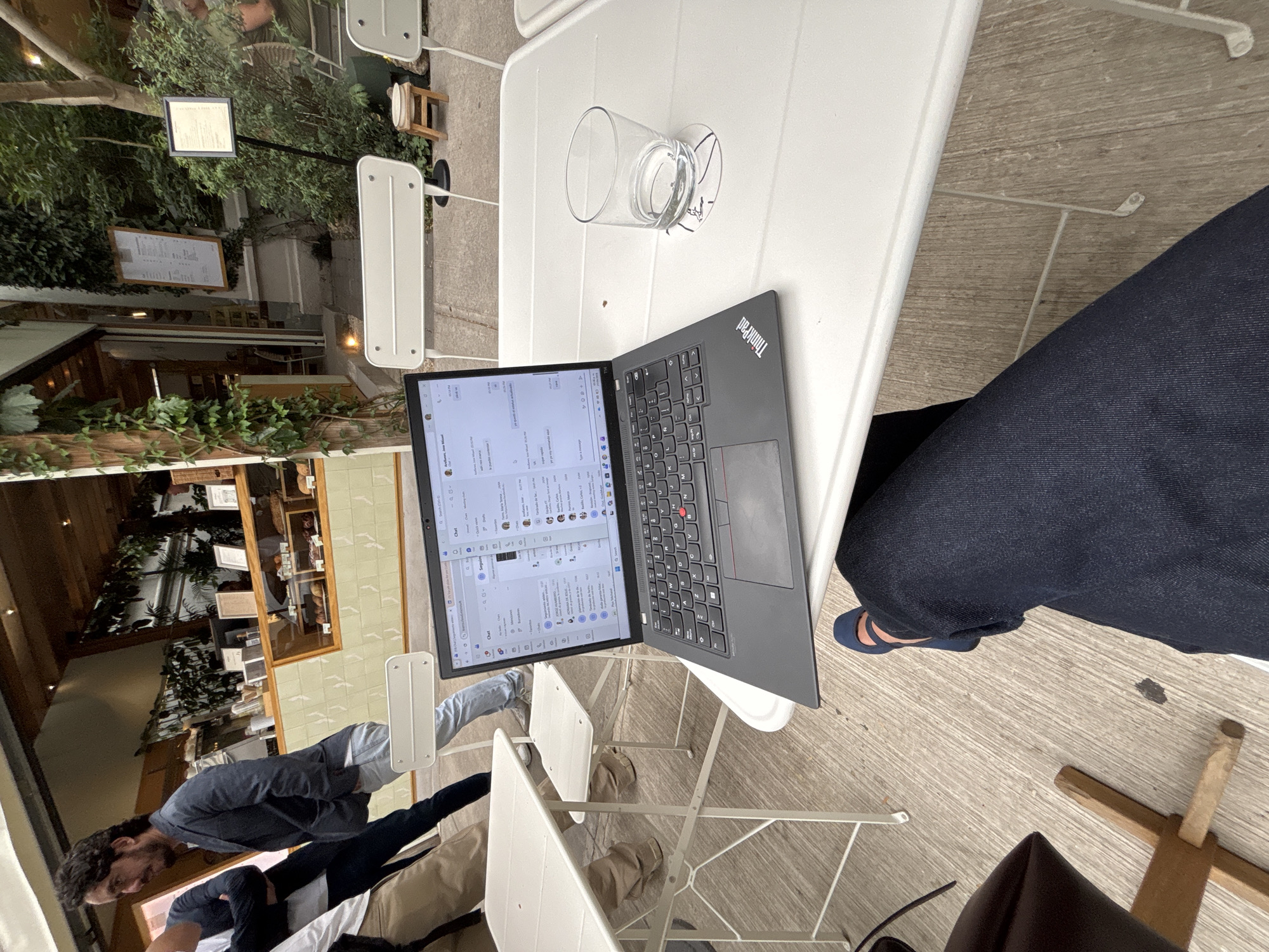 This image captures a daytime scene at an outdoor or semi-outdoor cafe or restaurant, likely in Mexico City. The main subject is a black Lenovo ThinkPad laptop open on a white metal folding table in the foreground. Beside the laptop, a clear glass with a small amount of liquid sits, leaving a wet ring on the table.
A person, whose lap and legs are visible wearing dark blue jeans and blue flat shoes, is seated at this table, seemingly using the laptop. The laptop screen displays what appears to be a messaging application, possibly Microsoft Teams or a similar client, with lists of contacts or channels and message previews. Visible Spanish text on the screen includes words like "Anuncios," "Saludos," "Hoy," "Semana," and "Escribir un mensaje." The "ThinkPad" logo is visible on the laptop's lid.
In the background, other white metal tables and chairs are scattered. Several people are present; one person wearing a blue shirt and light jeans stands to the left, partially obscured, facing away from the camera. Another person's head and shoulders are visible further back. The setting features lush greenery, including plants and trees, suggesting a pleasant, natural environment. Parts of a building with wooden frames and shelves, possibly showcasing baked goods or other items, are visible through the foliage, indicating the cafe's interior. Two signs, one black-framed and one wooden-framed, are posted amidst the greenery, likely displaying menus or information, though their text is unreadable. The flooring is a light-colored wooden or tiled surface. The overall atmosphere appears relaxed, suitable for working or socializing.