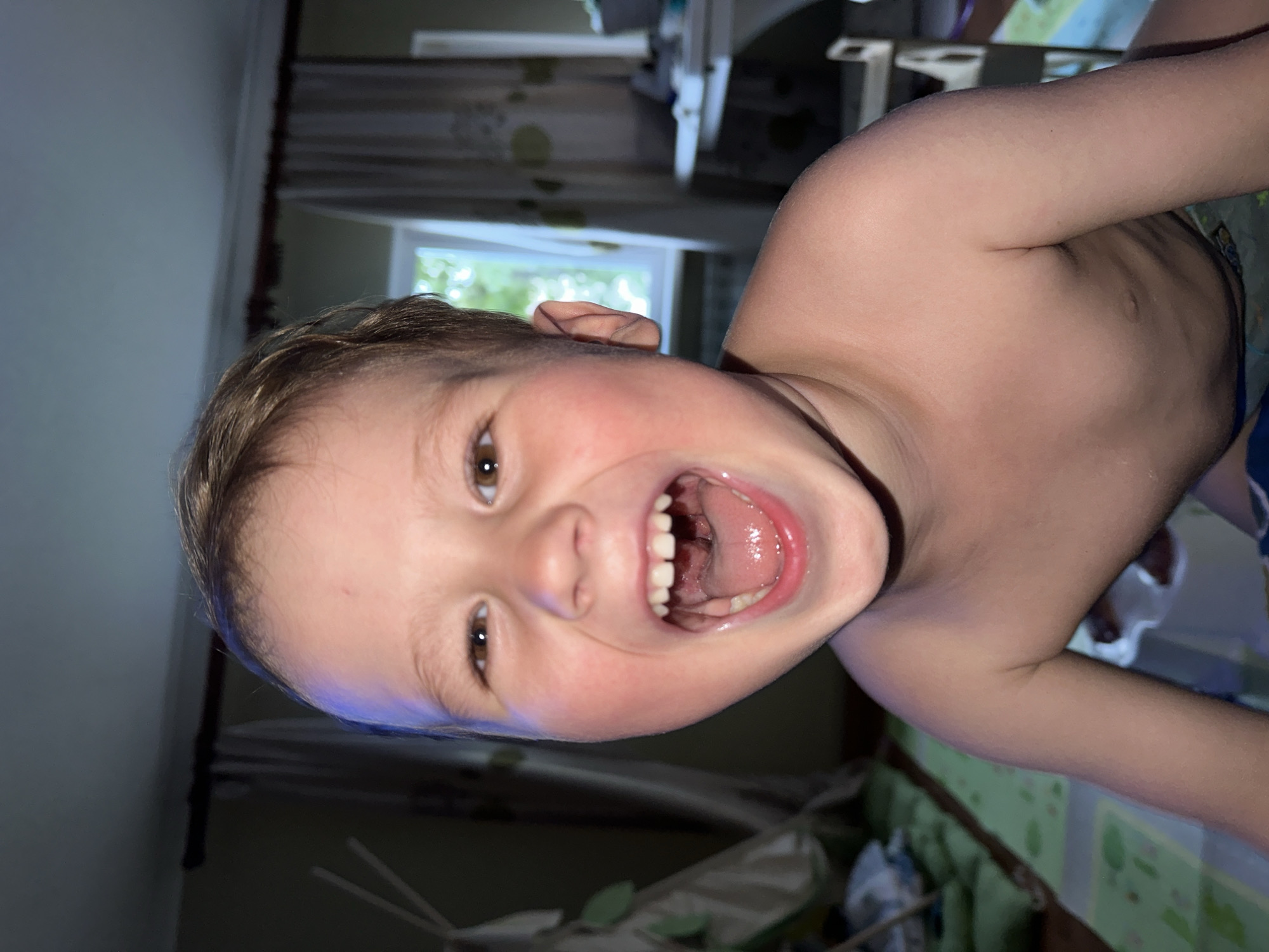 The image shows a close-up, waist-up, indoor portrait of a young, bare-chested Caucasian boy with light brown hair, taken from a slightly low angle. He is looking directly at the camera with a wide, open-mouthed expression of laughter or surprise, revealing his teeth and the inside of his mouth. His cheeks are flushed, and his eyes are open and bright. The background is dimly lit and appears to be a residential room. To the left, a portion of a teepee-like tent is visible, along with what looks like a green mat on the floor. In the center background, a window is visible with white curtains, and beyond the window, greenery suggests an outdoor environment. To the right, a table and chair are faintly discernible. The lighting creates a dramatic effect, with a bright light source from above and slightly to the left illuminating the boy's face. The overall impression is one of candid joy. The image's metadata, if available, would be needed to determine its specific location within Nizhny Novgorod, Russia.