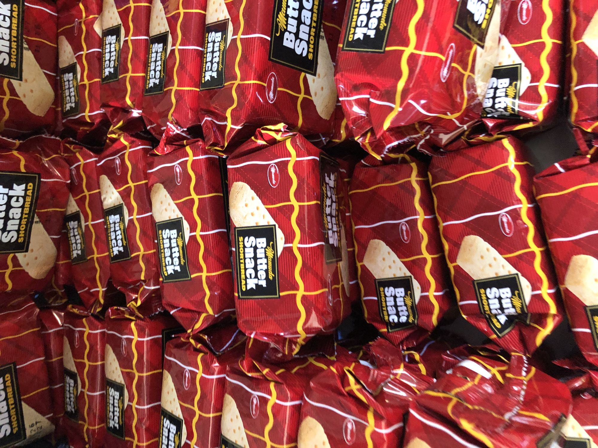 The image is a close-up, high-angle shot of multiple individually wrapped packages of "Mamuda Butter Snack Shortbread" biscuits. The packaging is predominantly red with a yellow and white checkered pattern and a gold wavy line design. Each package features a black label with white and gold text. The biscuits themselves are visible through some of the packaging, appearing light brown and rectangular with small perforations. The scene is likely an indoor retail environment, possibly a shop or market stall, given the arrangement of the stacked products. There are no people or specific identifiable landmarks visible in the image. The location is stated to be Bar Jirgi Summa, Nigeria, but this information cannot be visually confirmed from the provided image alone.