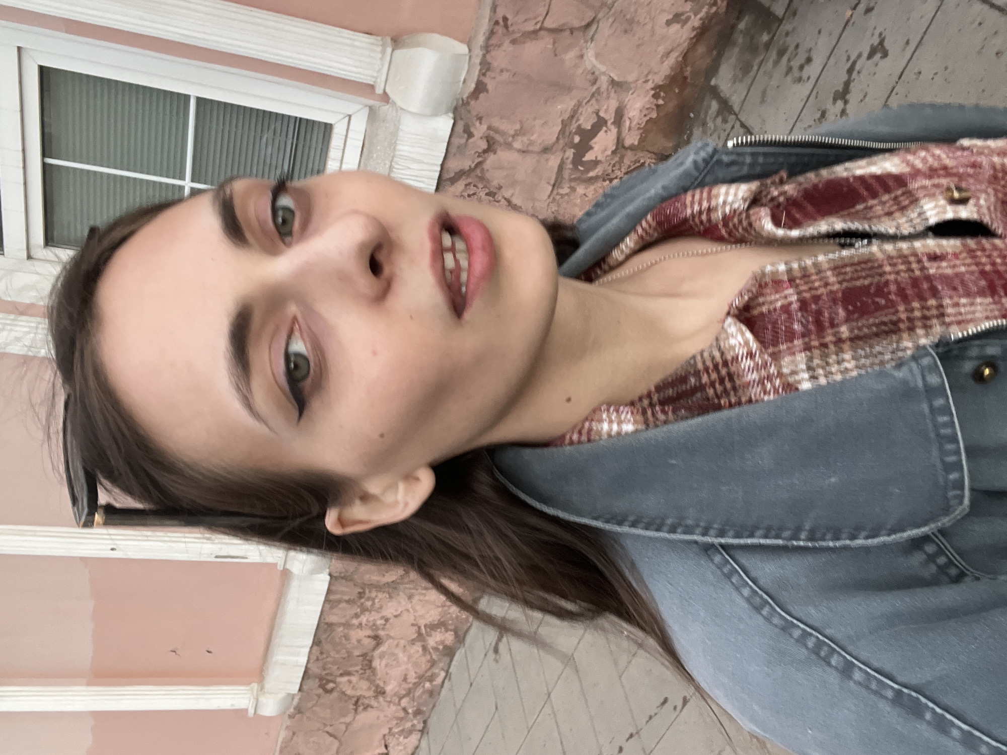 A selfie shows a woman in front of a building. She is wearing a grey denim jacket over a red and tan plaid shirt, with dark hair and green eyes. The background includes a pink building with white trim, a window with horizontal blinds, and a section of brick wall. The ground appears to be paved. The location may be Krasnoyarsk, Russia.