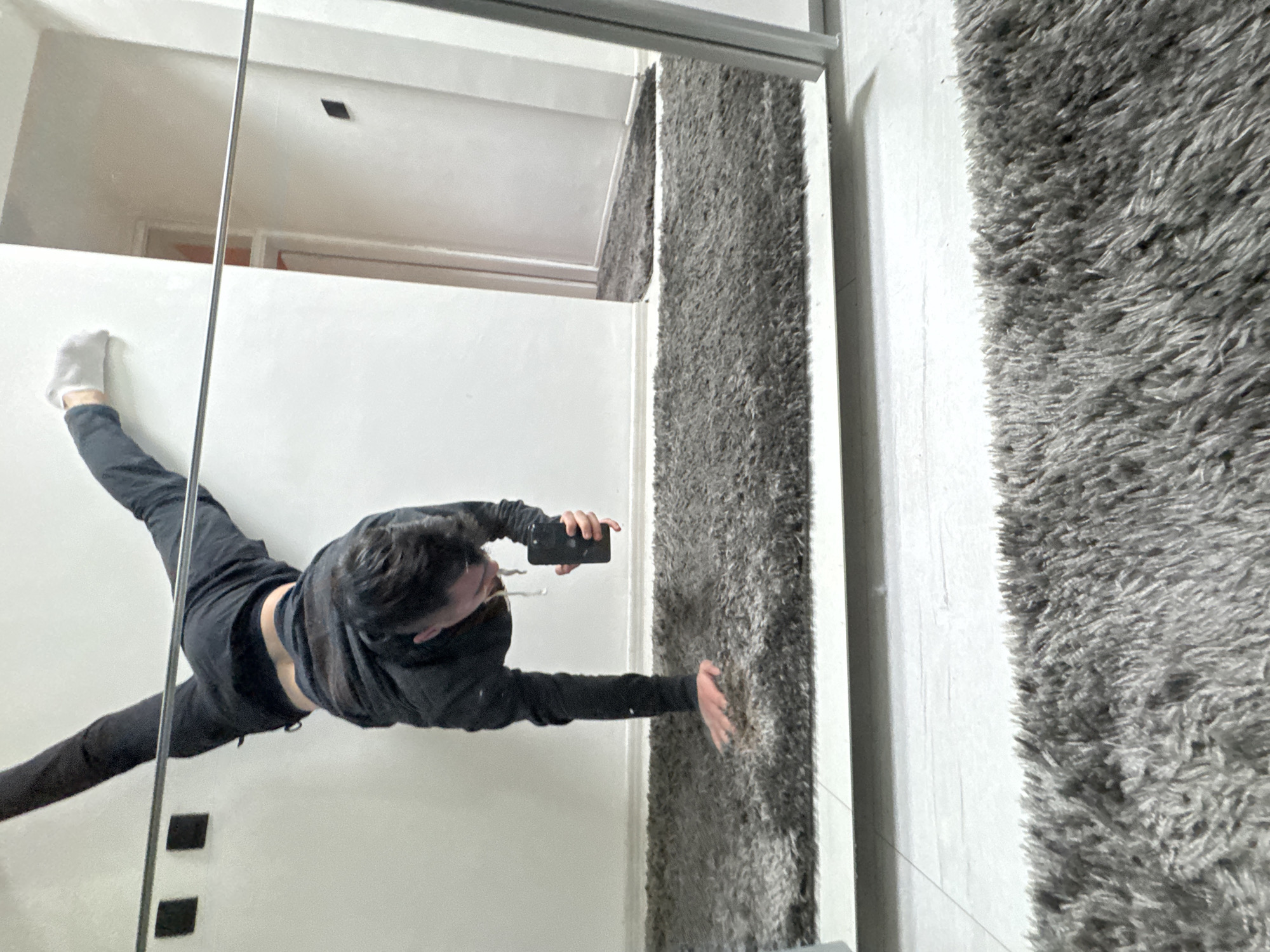 This media file captures a rotated mirror selfie taken indoors. A person, appearing to be male, is the primary subject, dressed in a black hoodie and black pants. They are holding a black smartphone in their right hand, using it to take the picture.
The person is striking a dynamic pose, with their left leg extended upwards and sideways, their foot (clad in a white sock) pressing against a light-colored wall. Their right arm is also extended, reaching towards a shaggy gray rug that covers what appears to be another wall in the reflection. The pose gives the impression that the person is doing a split or climbing on the wall. Their midriff is visible between their hoodie and pants.
The setting appears to be a modern indoor space. The mirror reflects a light-colored, possibly white, room with minimalist design. In the reflection behind the person's extended leg, two black square objects are visible on the wall. Above the person's head, the reflection shows a ceiling and what seems to be a doorframe or opening with an orange-ish interior. A thin, reflective metal bar runs vertically through the center of the image, potentially part of the mirror's frame or a room partition.
The lighting is soft and even, suggesting daytime conditions without direct harsh sunlight. There is no visible text in the image, nor are there any specific cues within the image itself to confirm the location as Sofia, Bulgaria. The overall impression is one of a playful or artistic self-portrait.