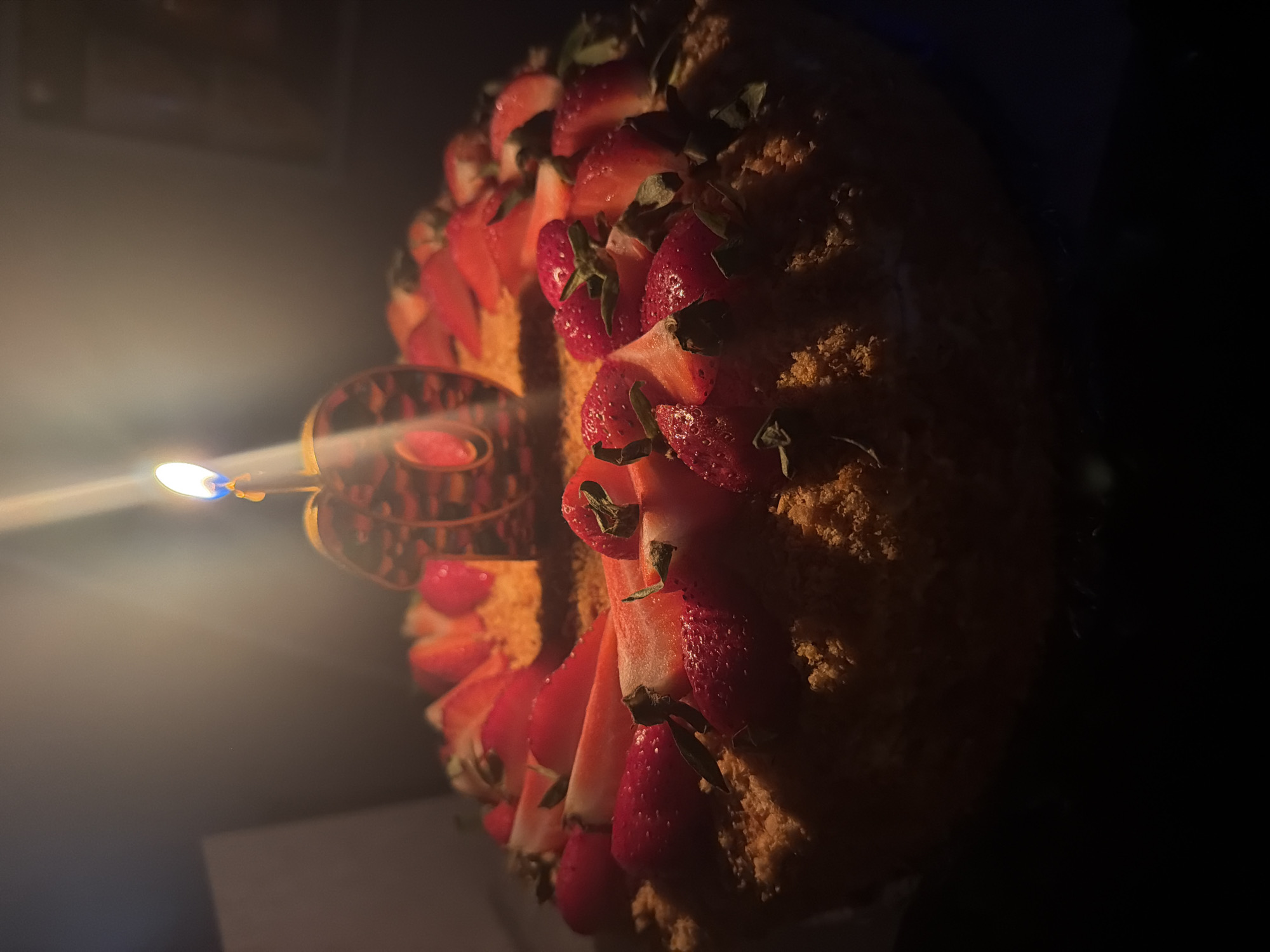 A birthday cake is decorated with sliced strawberries and a lit candle in the shape of the number "10". The cake is round and appears to be placed on a dark surface, possibly a table. The background is dark and out of focus, with a faint light source visible at the top left, suggesting an indoor setting. There are no people or other objects clearly visible. The image appears to be taken in Votkinsk, Russia, though no specific Votkinsk landmarks or identifying features are present in the image itself.