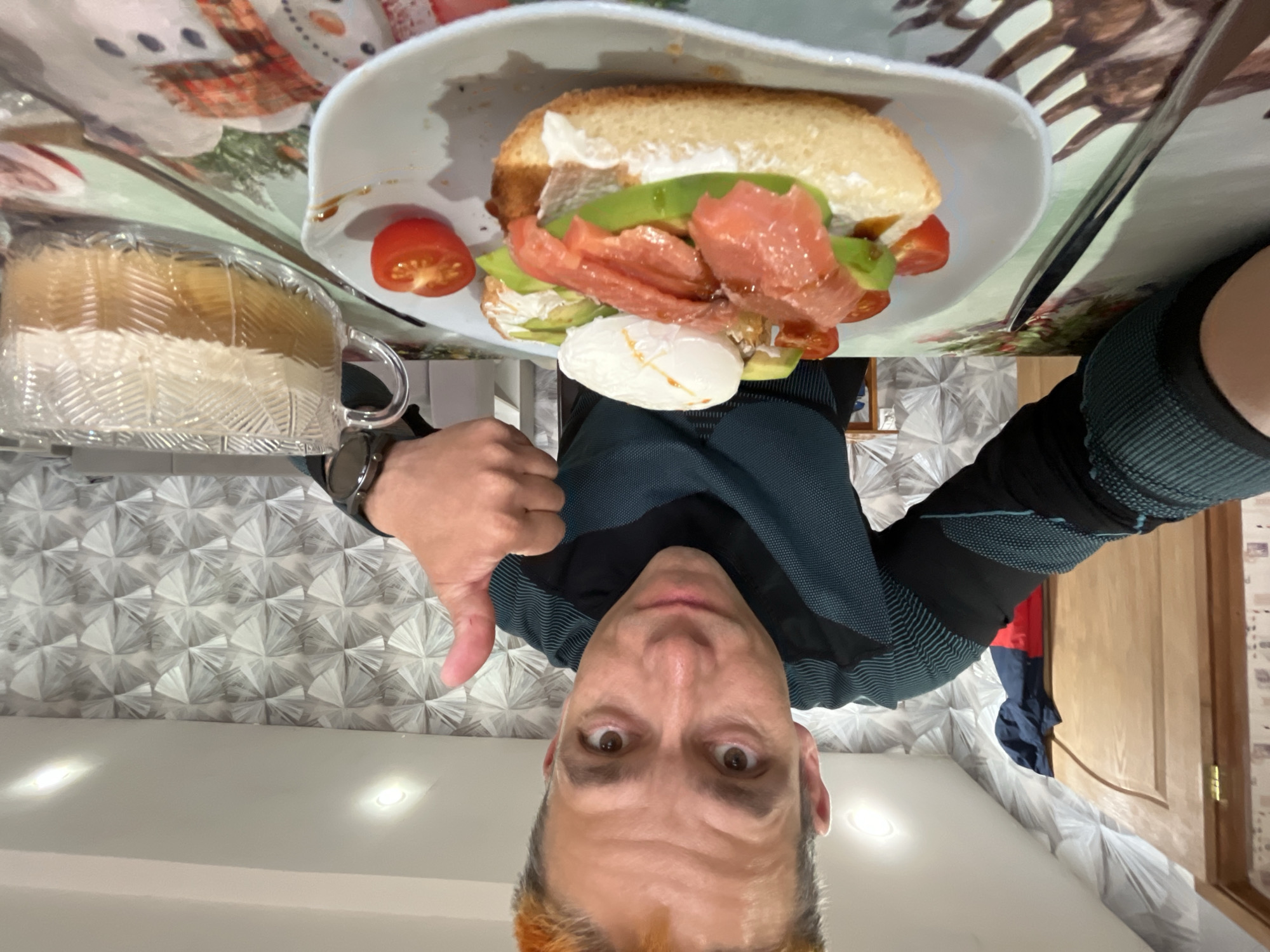 This image, rotated 90 degrees clockwise, depicts a man taking a close-up selfie while presenting a meal in an indoor setting in Chaikovsky, Russia. The man is positioned centrally, looking directly at the camera with wide, expressive eyes and giving a thumbs-down gesture with his right hand. He has short hair with visible reddish-orange highlights and is wearing a dark, form-fitting long-sleeved top with a textured pattern. A smartwatch is visible on his left wrist.
In front of him, on a white plate, is an elaborate open-faced sandwich or toast. It features a substantial piece of bread or bun spread with a creamy white topping, layered with green avocado slices, thin strips of what appears to be smoked salmon or cured fish, and several halved cherry tomatoes. A perfectly formed poached egg or a ball of burrata cheese is placed alongside the main ingredients. Next to the plate is a clear, patterned glass mug containing a light-colored beverage, possibly coffee or tea with milk.
The meal is placed on a table covered by a decorative placemat with a winter or Christmas theme, showing illustrations of deer and snowmen. The background reveals a wall with a modern, geometric pattern and a light-colored wooden door. Recessed lights are visible in the ceiling above. The man's wide-eyed expression and thumbs-down gesture convey a humorous or critical reaction to the food. There is no visible text in the image.
