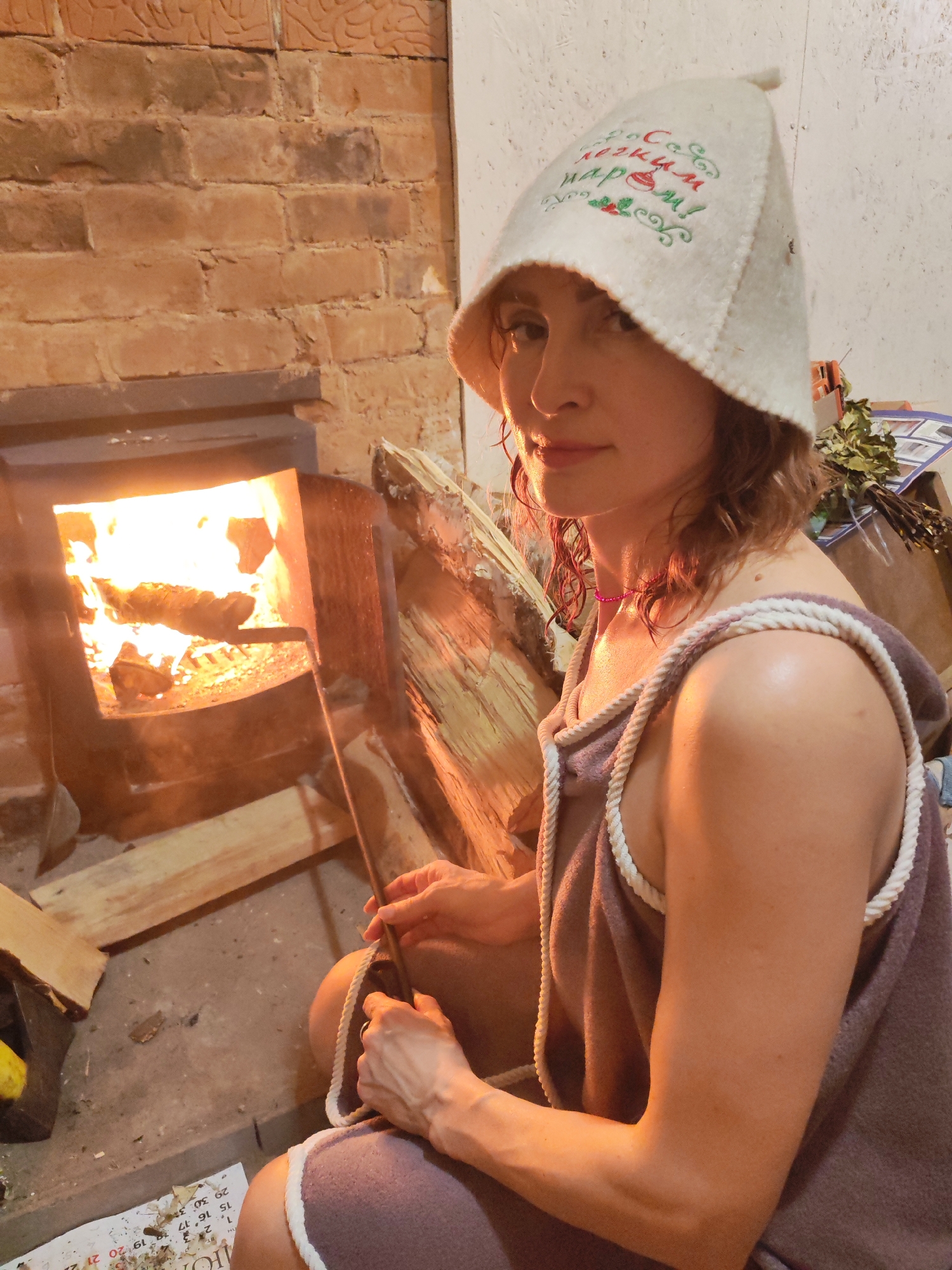 A woman is sitting near a wood-burning stove with a fire burning inside. She is wearing a white, felt hat with embroidered text and images, and a mauve-colored towel wrap with white rope trim around the edges. Her wet hair is visible around her face. She is holding a metal poker and appears to be tending to the fire. The stove is set into a brick wall. A stack of wood is adjacent to the stove. The location appears to be indoors, possibly in a sauna or bathhouse. A calendar on the floor near her feet. A bundle of leafy twigs is behind her. The city and country are unknown.