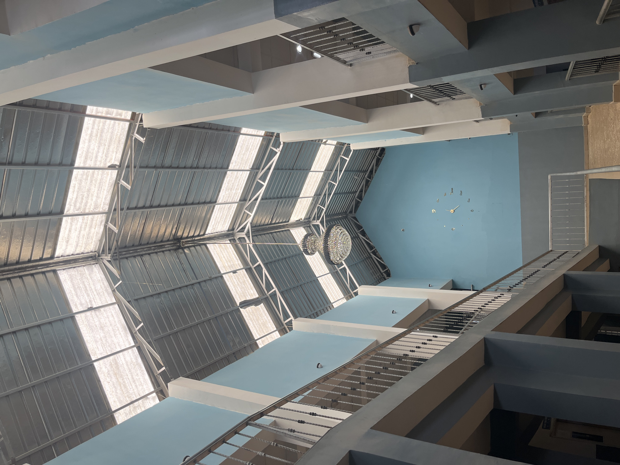 The image depicts the interior of a multi-level building with a high, open ceiling structure in Jos, Nigeria. Natural light streams in through a translucent roof composed of corrugated sheeting supported by metal trusses. The walls are painted in alternating shades of light blue and grey, with white vertical support columns.
On the left and right sides of the frame, elevated walkways or balconies with metal railings are visible, suggesting a circulation space. A large, ornate chandelier hangs from the ceiling in the center of the image. On the back wall, a modern, minimalist clock with thin hands is mounted.
The overall impression is one of a spacious, modern interior, possibly a public building like a school, shopping center, or administrative complex, designed to maximize natural light. The absence of people suggests the image was taken during a quiet period. The architectural style and the clock are contemporary.