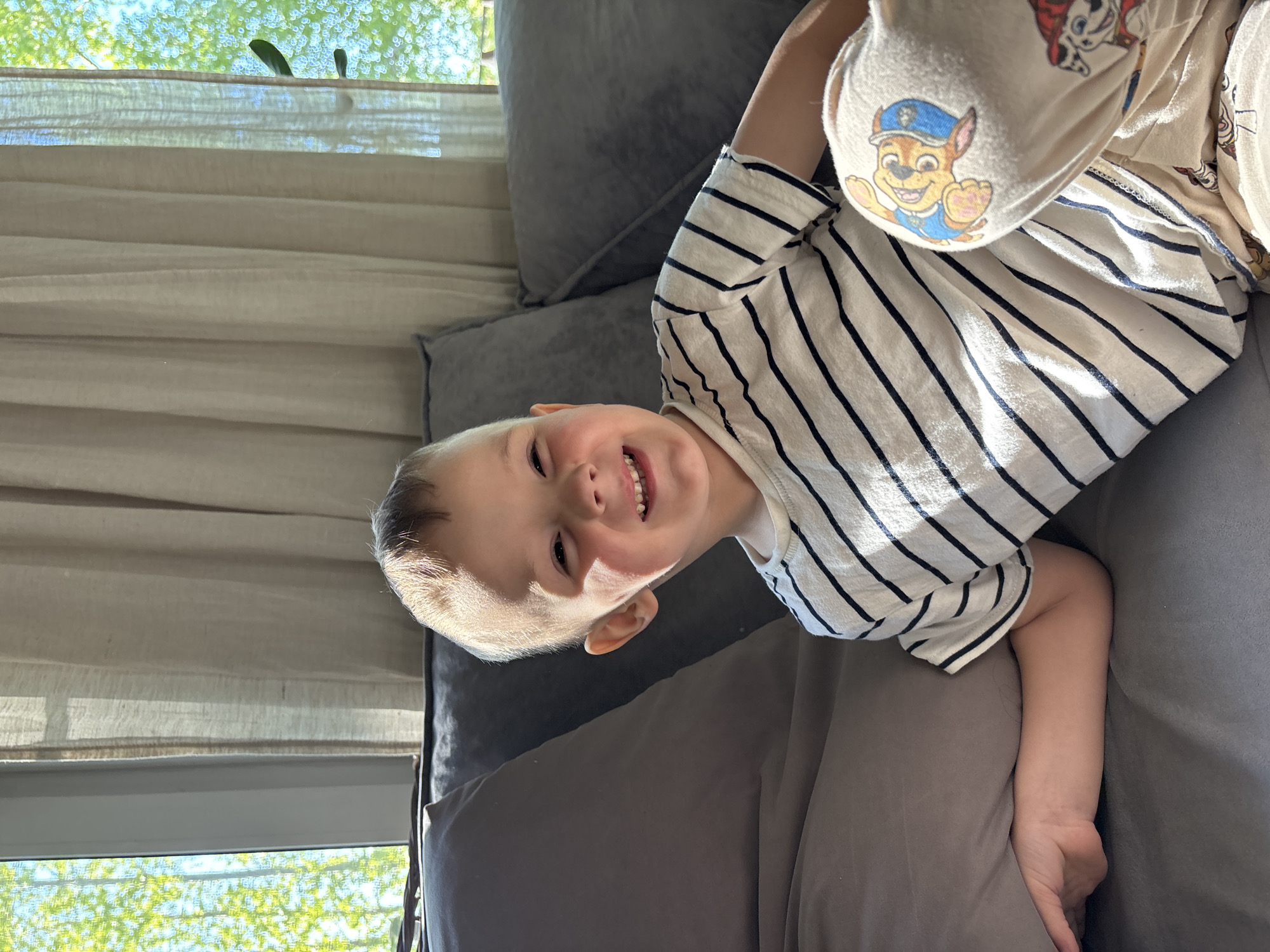 A young child is lying on a gray couch. The child has fair skin, short blond hair, and is smiling. They are wearing a white and black striped shirt and light-colored pants with a Paw Patrol design. One of the child's legs is bent upwards, revealing a Paw Patrol character on the sole of the child's slipper. The couch is comprised of gray cushions, and there are windows in the background with sheer curtains. Sunlight is visible through the window, and trees can be seen outside. The image was taken indoors. There is no indication of a location in Chaikovsky, Russia.