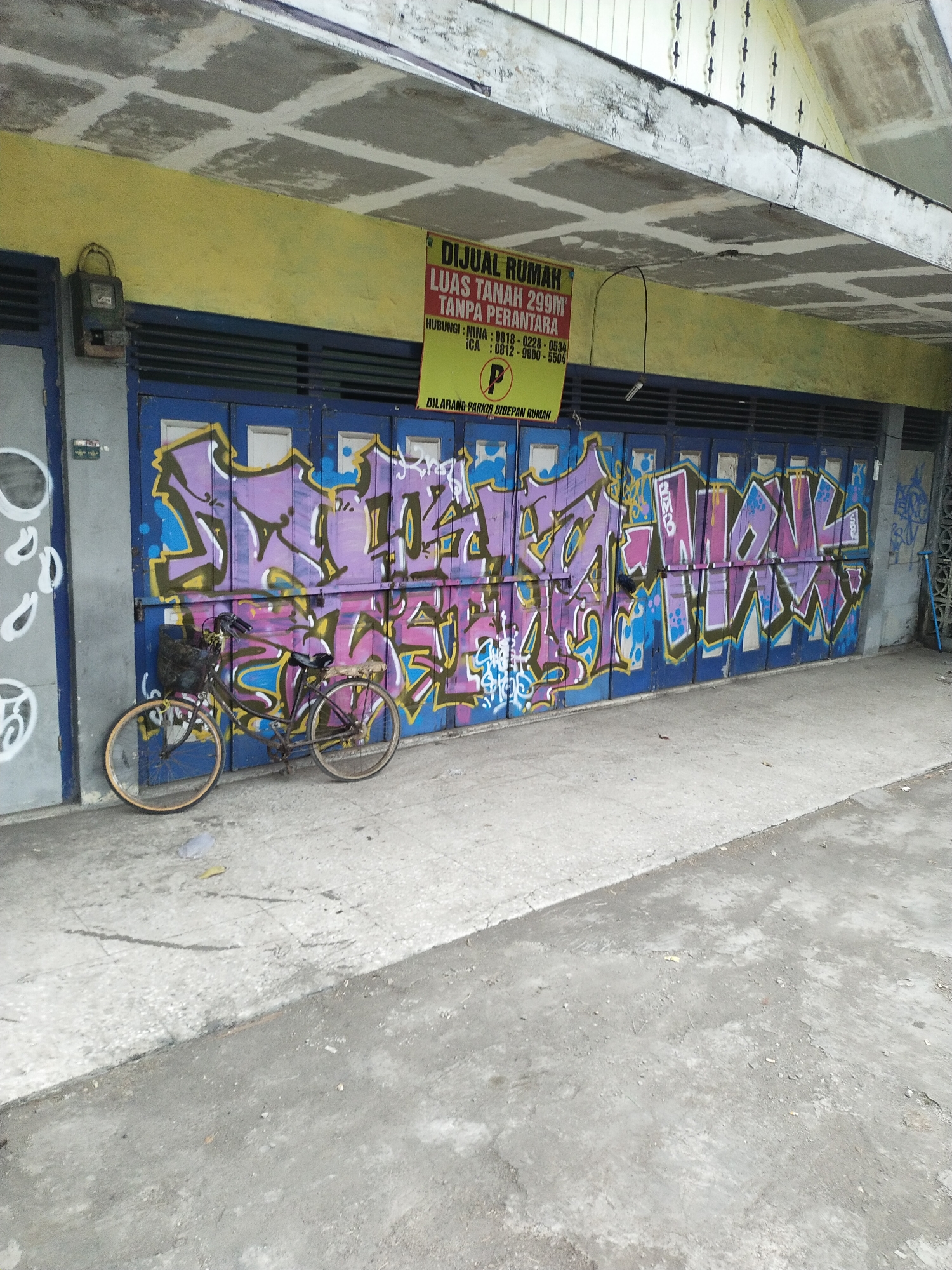 A weathered building facade in Kemlakagede, Indonesia, features blue roll-up doors covered in vibrant purple and yellow graffiti. A faded yellow sign above the doors advertises "DIJUAL RUMAH" (House for Sale) and lists property details and contact numbers. To the left of the graffiti-covered doors, a light gray door or wall has abstract white spray-painted circles. In front of the graffiti, a rusty bicycle with a black basket is parked on a concrete sidewalk. The sidewalk appears to be made of light-colored concrete with darker, uneven patches. Beyond the sidewalk is a rougher, gray concrete or asphalt surface. The ceiling above the facade is made of concrete panels with visible grid lines.