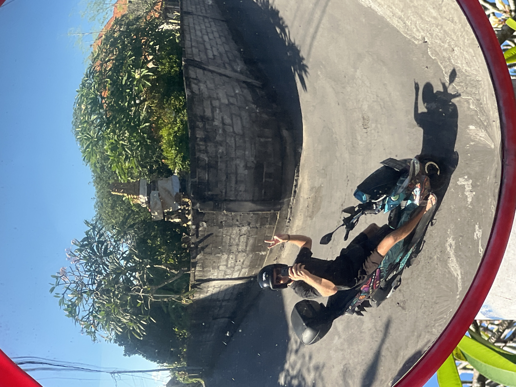 A person wearing a black t-shirt, black shorts, a black helmet, and sunglasses rides a blue and white patterned scooter. The rider is holding a smartphone in their right hand and making a peace sign with their left hand. The scooter has a black storage box on the back. The scene is reflected in a large, red-rimmed convex mirror, which distorts the view. Behind the scooter, a grey brick wall curves around a corner. Beyond the wall, lush green trees and vegetation are visible, with a hint of red-tiled roofs peeking through. The sky is clear and blue. The image appears to have been taken in a tropical or subtropical environment, likely Sukawati, Indonesia, given the architectural style and vegetation.
