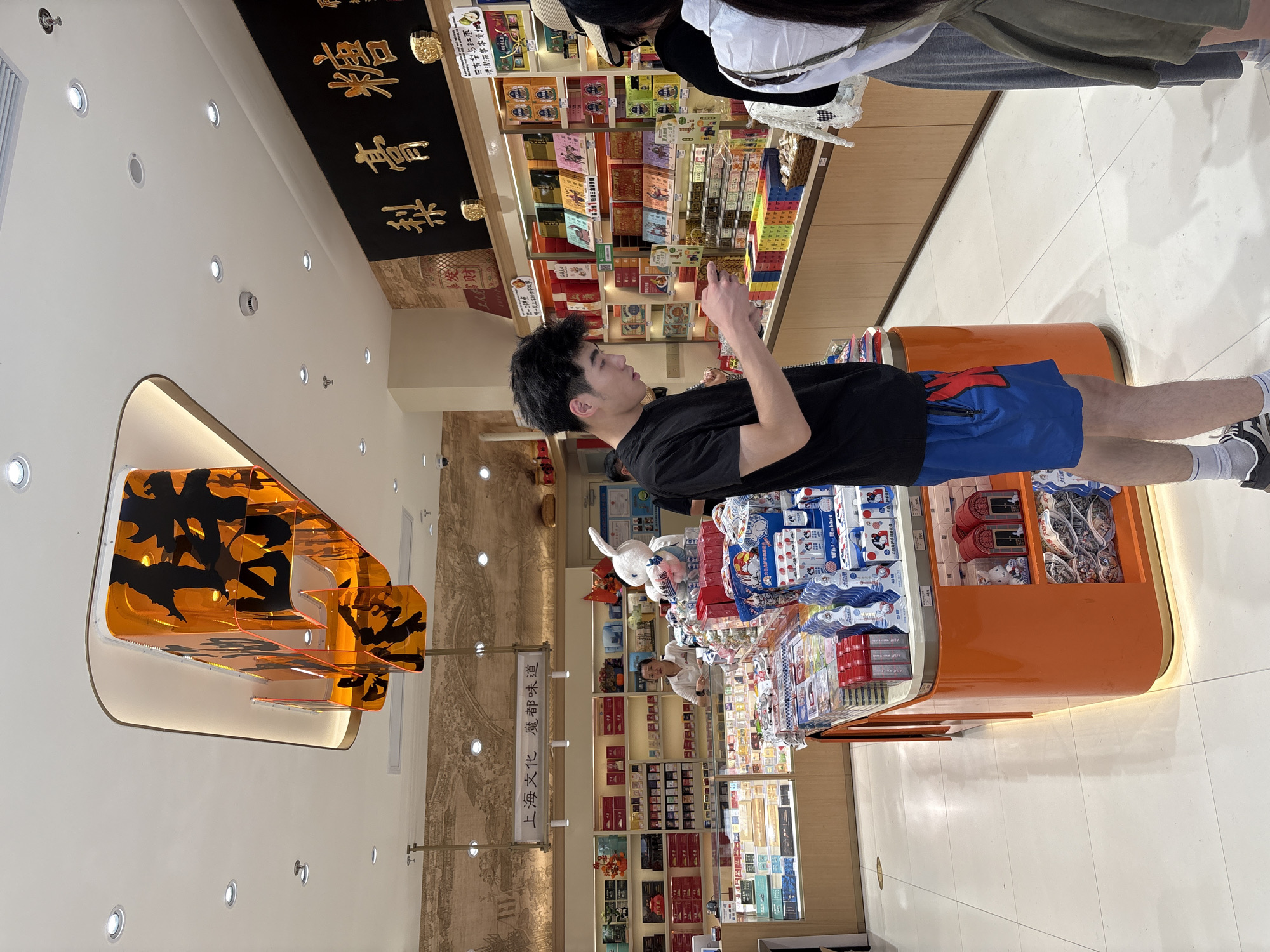 The image shows the interior of a shop in Shanghai, China, likely a confectionery or gift shop. A young man in a black t-shirt and blue shorts stands in the foreground, looking at his phone. Behind him, a woman with a white shirt is visible behind a counter. The shop is filled with shelves of products, many of which appear to be snacks or candies, displayed in boxes and packages. A large, orange display fixture with black Chinese calligraphy is suspended from the ceiling. The walls are decorated with a mural depicting a historical scene and Chinese characters. The floor is tiled in white. The overall impression is of a well-lit, modern retail environment selling traditional Chinese goods.