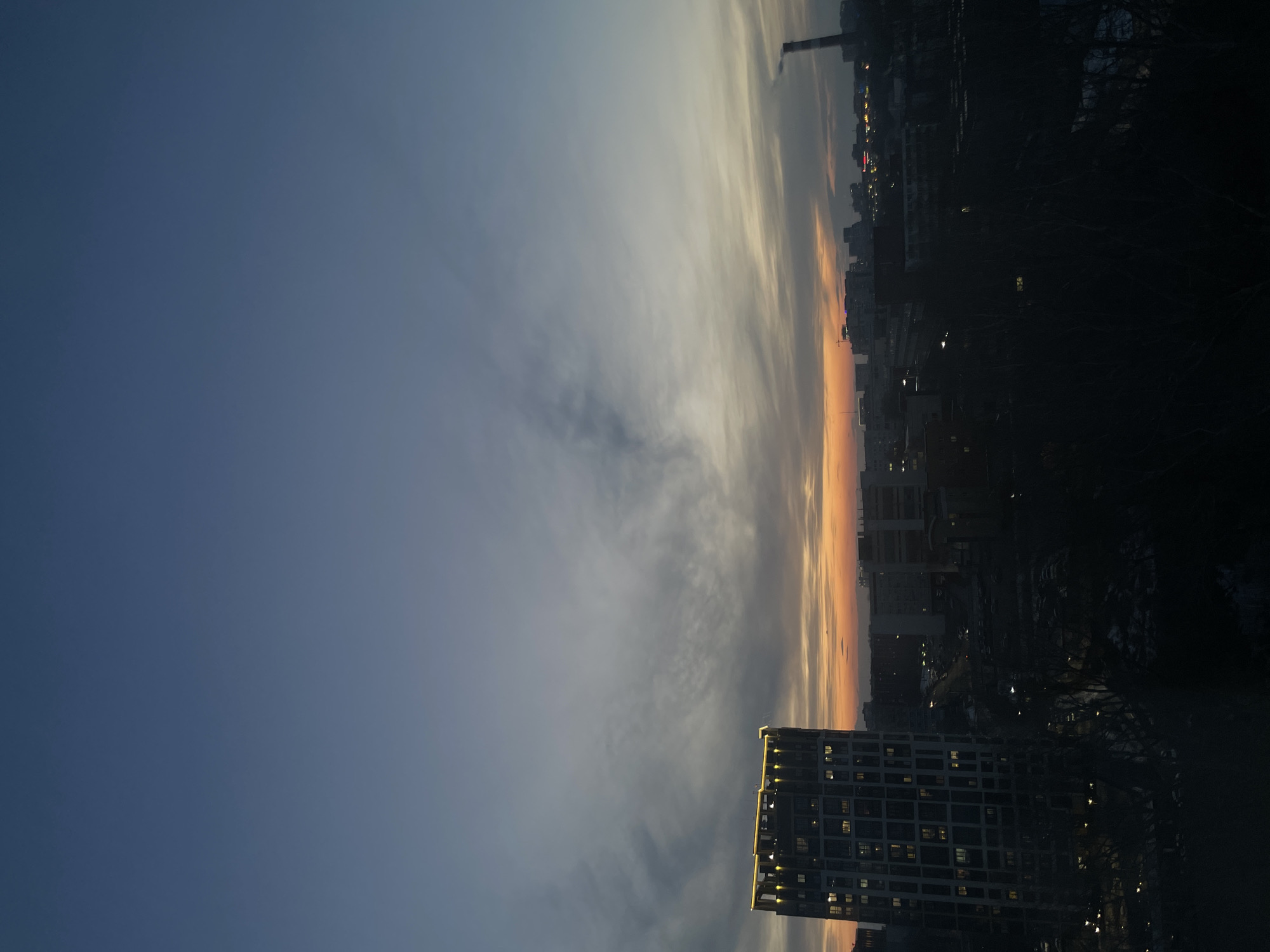 The image shows a dusk or dawn scene in what is likely Izhevsk, Russia, based on the provided context. In the foreground is a tall, modern apartment building with many lit windows. Above and to the left of the building, the sky transitions from deep blue to a lighter, cloudier horizon with streaks of orange from the setting or rising sun. Further in the distance, a darker cityscape is visible, suggesting a densely populated urban area. The scene is likely captured from a high vantage point, giving a broad view of the city. There are no people visible in the image.