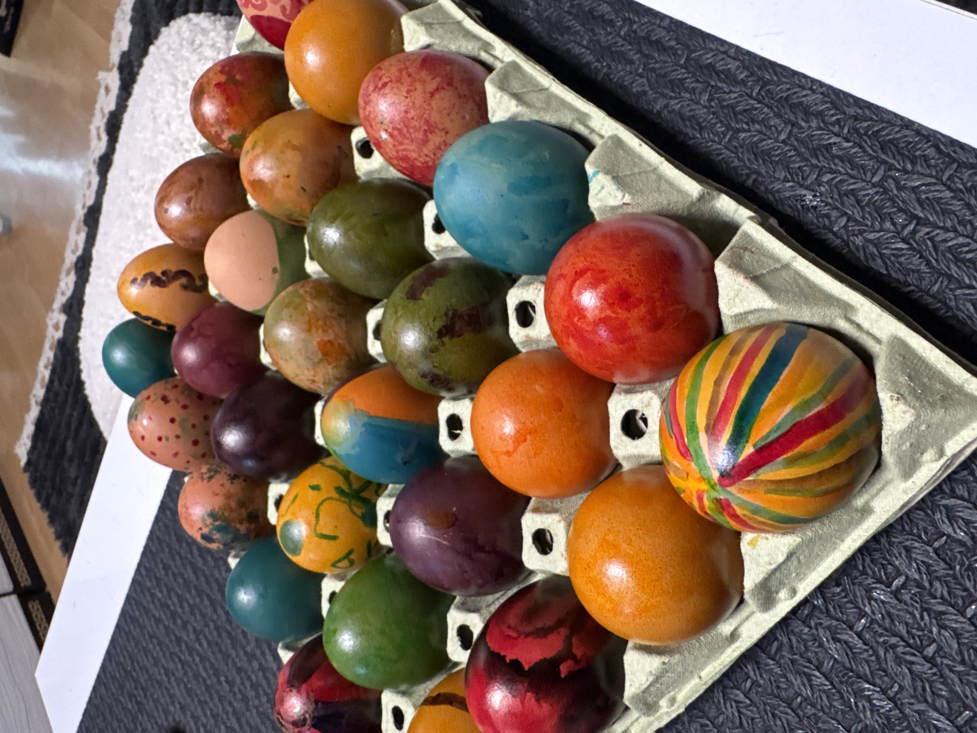 A high-angle shot displays a carton of colored eggs. The eggs feature various designs, including solid colors (orange, red, blue, green, purple), speckled patterns, and one with painted stripes. The carton sits on a surface with a textured grey fabric. In the background, a white surface and a patterned carpet are visible. The location is unidentifiable as Sofia, Bulgaria, from this image.
