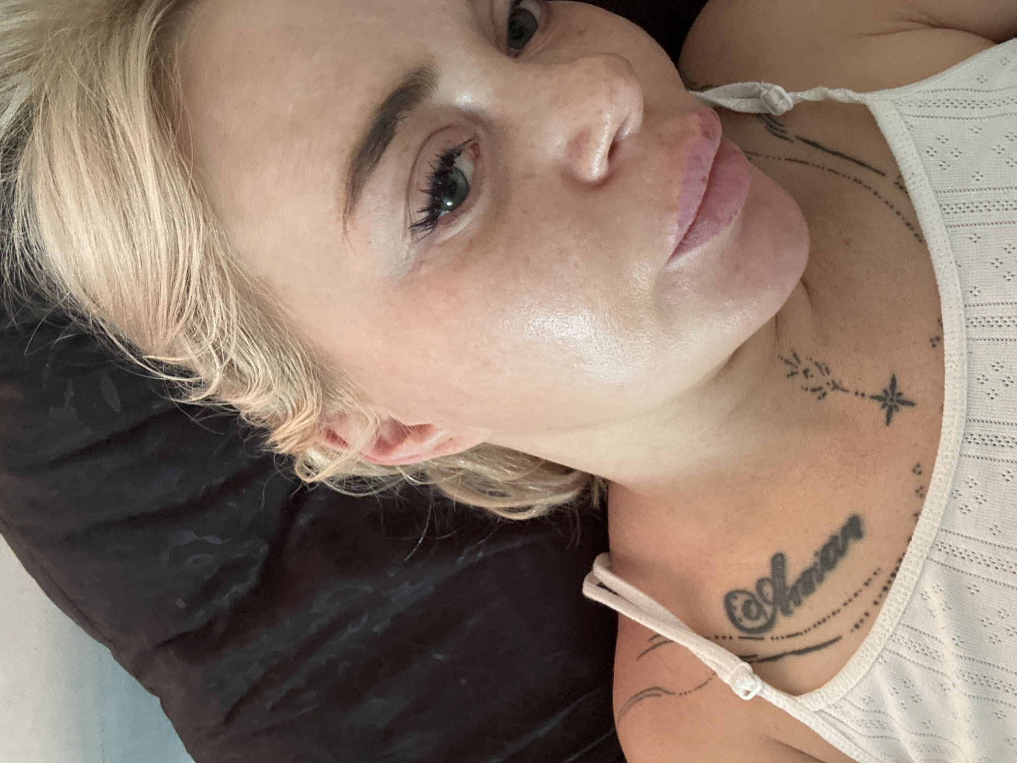 A close-up, overhead shot shows a blonde woman with fair skin and visible freckles lying down. Her hair is pulled back loosely, with some strands framing her face. She is wearing a white tank top with thin straps. Tattoos are visible on her upper chest and left shoulder. The background consists of a dark, textured surface. The image is lit from above, highlighting the woman's facial features and skin texture.