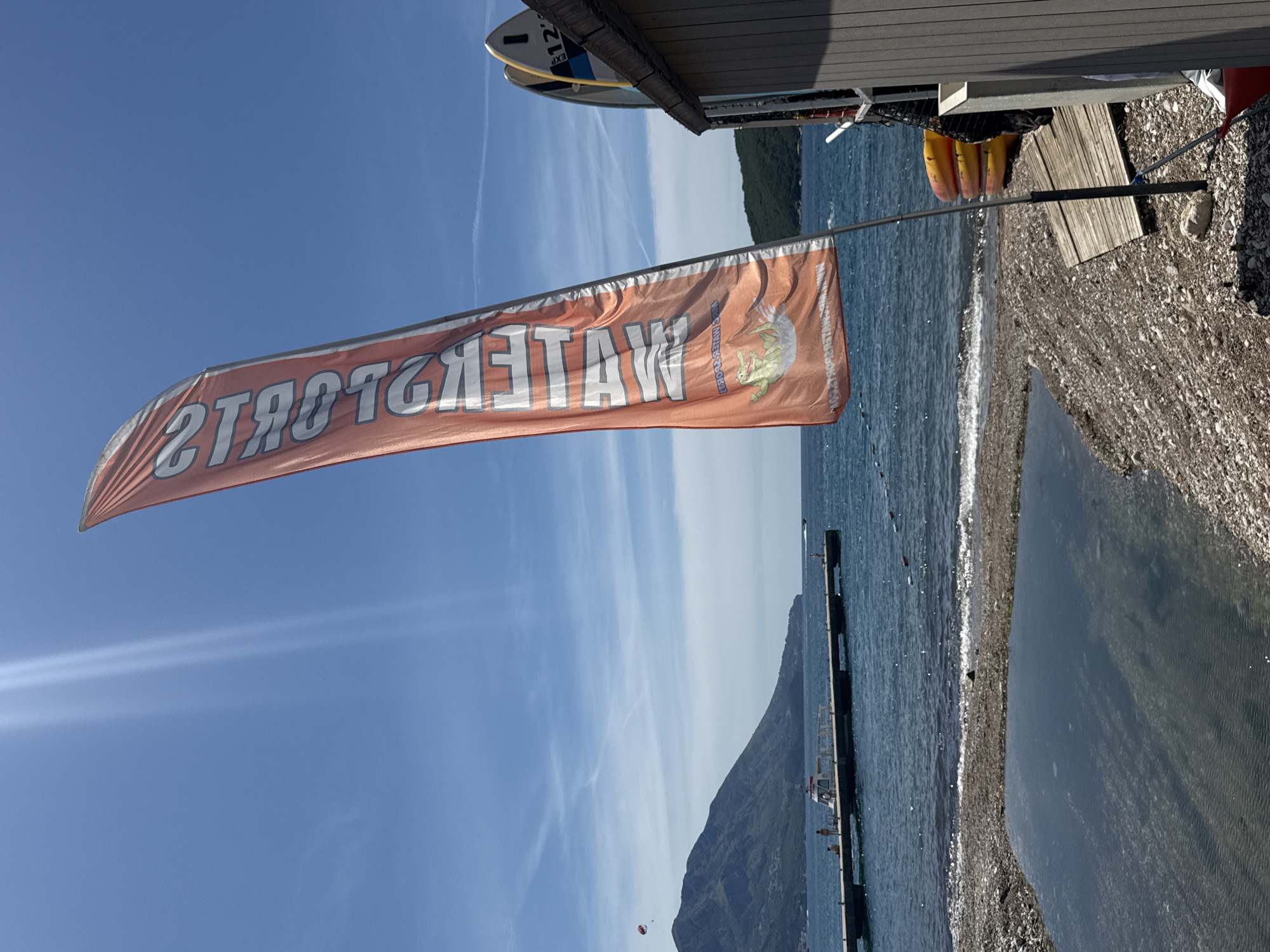 An orange banner with white lettering stands tall on a pole on a rocky beach in Budva, Montenegro. The banner reads "WATERSPORTS" vertically. A cartoon crocodile is depicted on the lower part of the banner, in a splash. Behind the banner, the deep blue Adriatic Sea stretches out, with a long pier in the distance. On the pier, several people are visible, as well as a white boat. Further in the distance, a large, green mountain looms under a clear blue sky with wispy clouds. To the right of the banner, a wooden structure is partially visible, with surfboards leaning against it. In front of the structure are several yellow kayaks stacked on top of each other. The beach itself is covered in small, grey pebbles and features a small, shallow pool of water reflecting the sky.