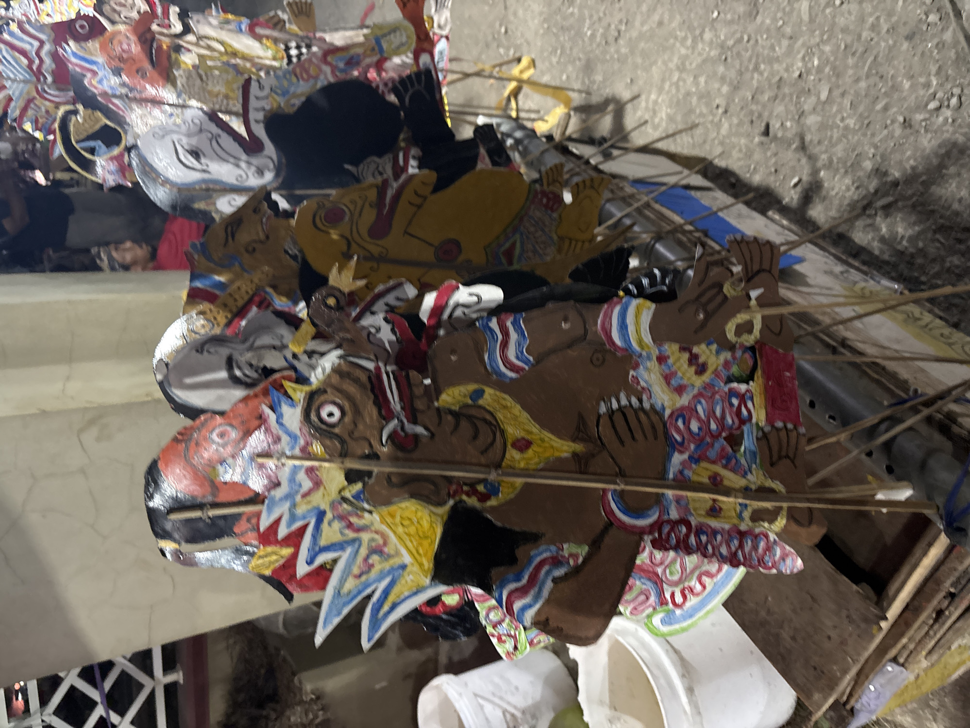This is a medium shot, eye-level, vertical photograph of a collection of Wayang Kulit puppets displayed on a table or stall. The puppets are made of leather and are ornately decorated with paint in various colors, including red, black, white, yellow, blue, and pink. They are mounted on sticks for manipulation. The puppets depict various characters, some with elaborate headdresses and facial features. In the background, out of focus, are some white buckets and a person wearing a red shirt. The ground appears to be dirt or gravel. The overall scene suggests an outdoor market or cultural display. The location is identified as Panongan, Indonesia.