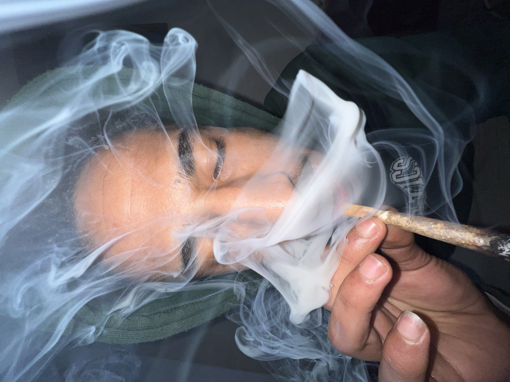 An individual with medium-dark skin, wearing a dark green hooded garment, is depicted in a close-up, dimly lit scene. The subject's eyes are closed, and their face, including eyebrows, nose, and mouth, is partially obscured by dense, white smoke. The right hand holds a lit, hand-rolled cigarette, likely a joint, between the thumb and index finger. The cigarette's tip shows active embers, and a significant volume of smoke emanates from it, swirling around the subject's face and hand. A faint, stylized logo, possibly "JCS" or "ACS", is visible on the subject's clothing below the hand, partially covered by smoke. The background is dark and indistinct. The specific location within Tanger, Morocco, is not discernible from the visual information presented in the image.