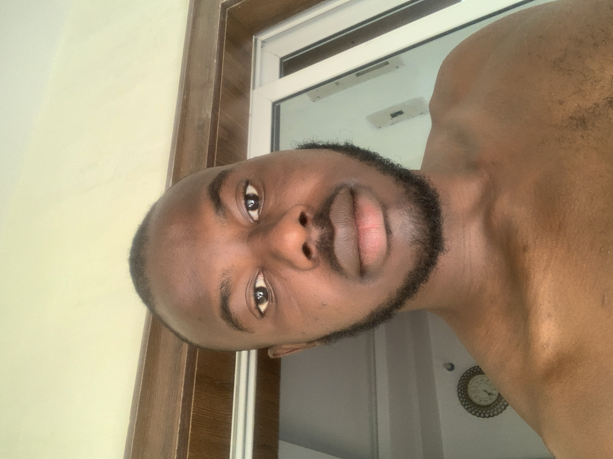 A man with dark skin, a closely shaved head, and a short, well-groomed beard is positioned close to the camera. The man's face is the primary subject, focusing on his eyes, nose, and mouth. The background appears to be an indoor setting, potentially a room with a window frame partially visible. A clock is on the wall. The location is Jos, Nigeria. The man is shirtless.