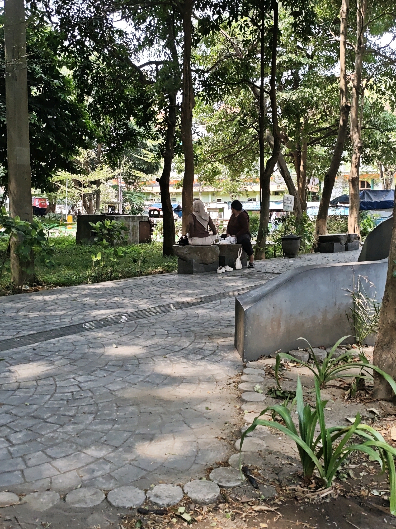 The image shows a park scene in Kediri City, Indonesia. In the mid-ground, two individuals are seated on a stone bench. One person is wearing a brown hijab and a brown top, and the other is wearing a brown long-sleeved top and dark pants. Between them on the bench are white objects, possibly bags or food containers. To the left of the bench, a white plastic bag and a pair of white slip-on shoes are visible on the ground. The individuals are facing each other. The environment consists of trees with dense foliage providing shade, a paved pathway made of grey interlocking bricks, and some low-lying greenery and plants. In the background, buildings with orange and yellow accents are visible, suggesting a residential or commercial area adjacent to the park. The ground in the foreground is also paved with similar grey bricks arranged in a circular pattern.
