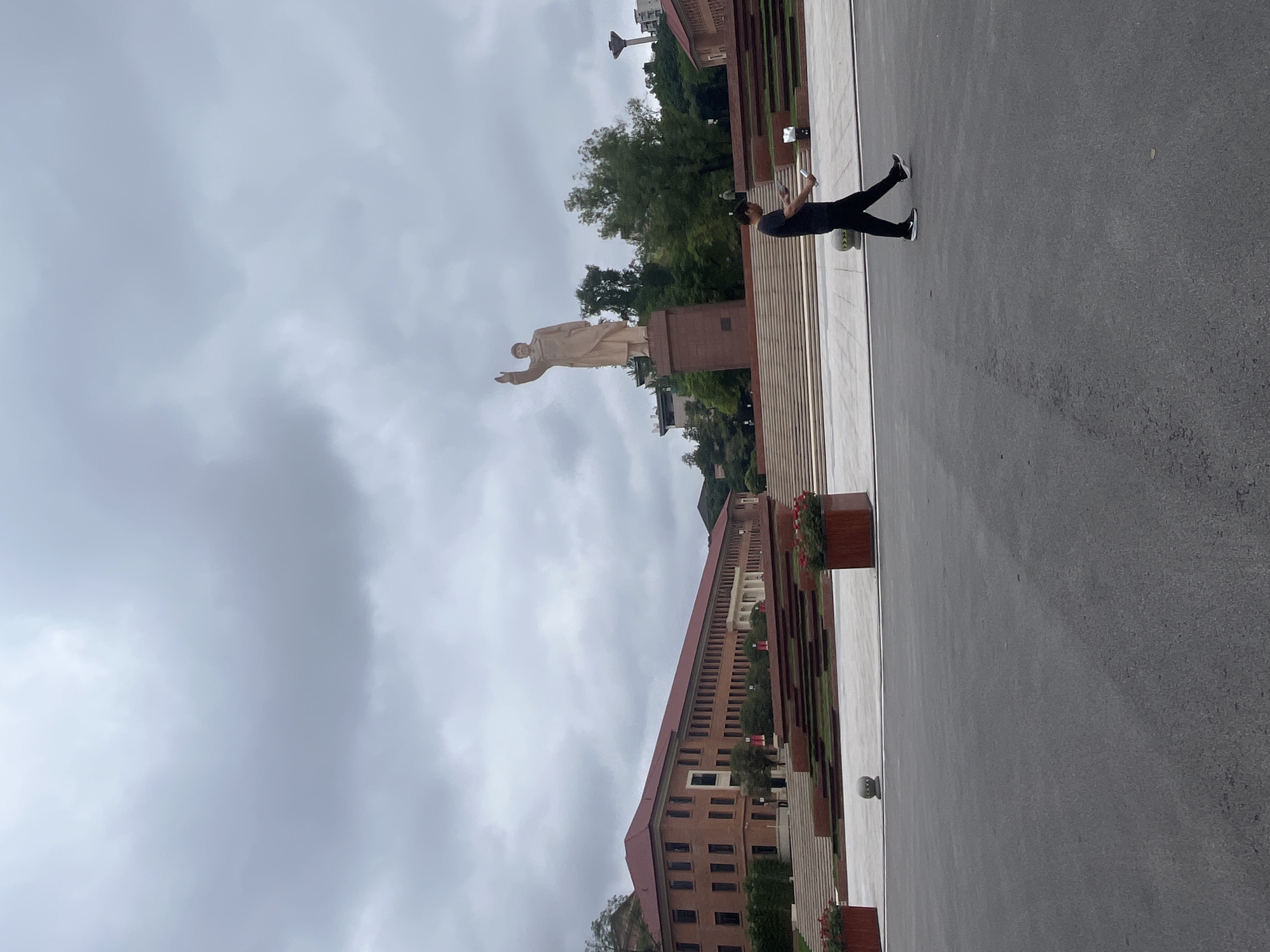 The image shows a man walking in a plaza in front of a large building and a statue in Yuzhong District, China. The man is wearing a black t-shirt and black pants, and he is holding a white object in his hands. The building is a red brick structure with a long facade, and it appears to be a school or government building. The statue is of a man in a suit, and he is raising his right hand. The sky is cloudy, and the overall lighting is overcast. There are also trees and plants in the background.