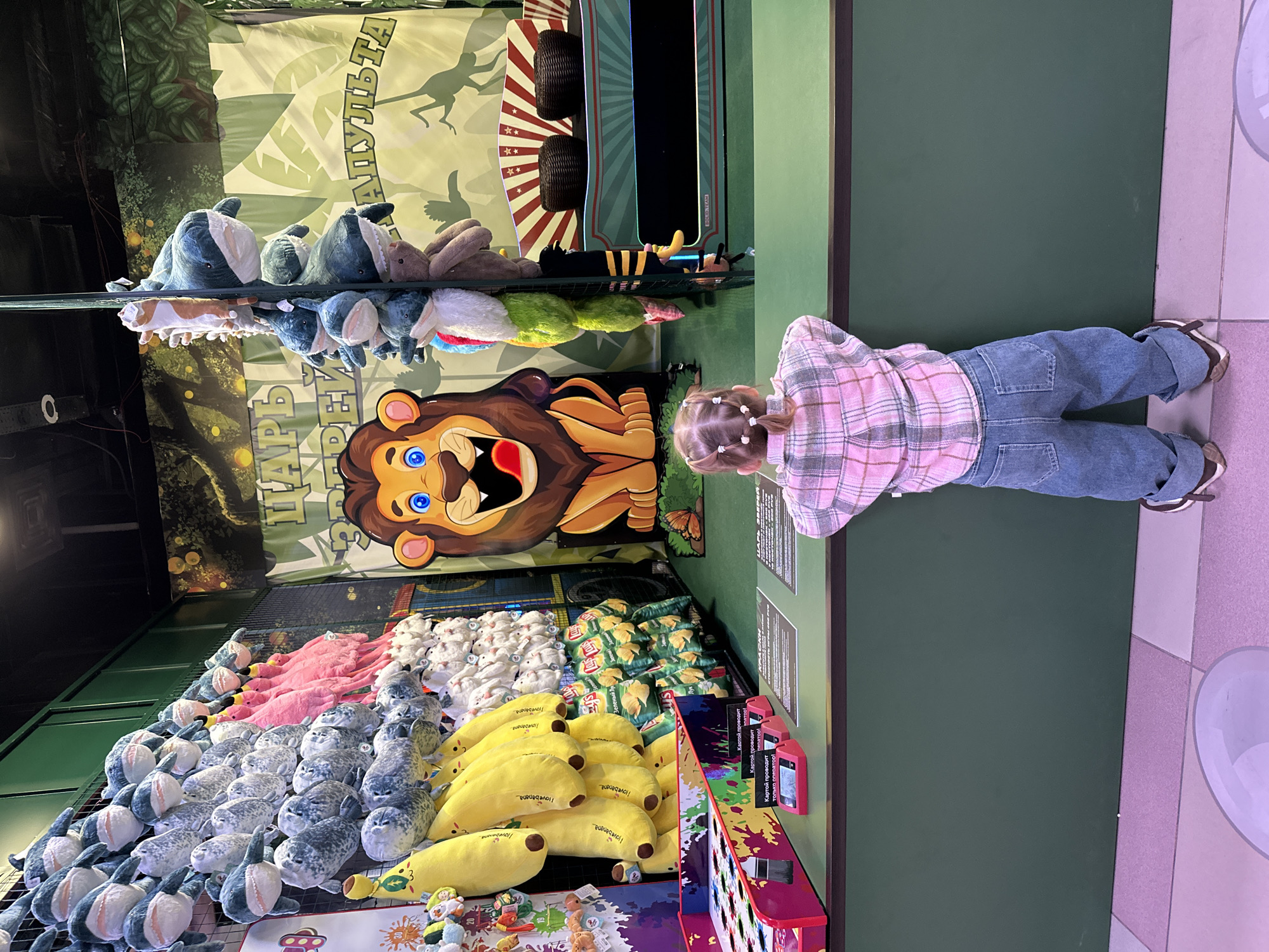 A young child stands next to a game called "Tsar Apult", likely in a shopping mall in Yekaterinburg, Russia, as seen in the background. The game features several shelves of stuffed animals, including blue seals, pink flamingos, white rabbits, yellow bananas, and bags of Lay's potato chips as prizes. The game also has a cartoon lion target for players to hit.
