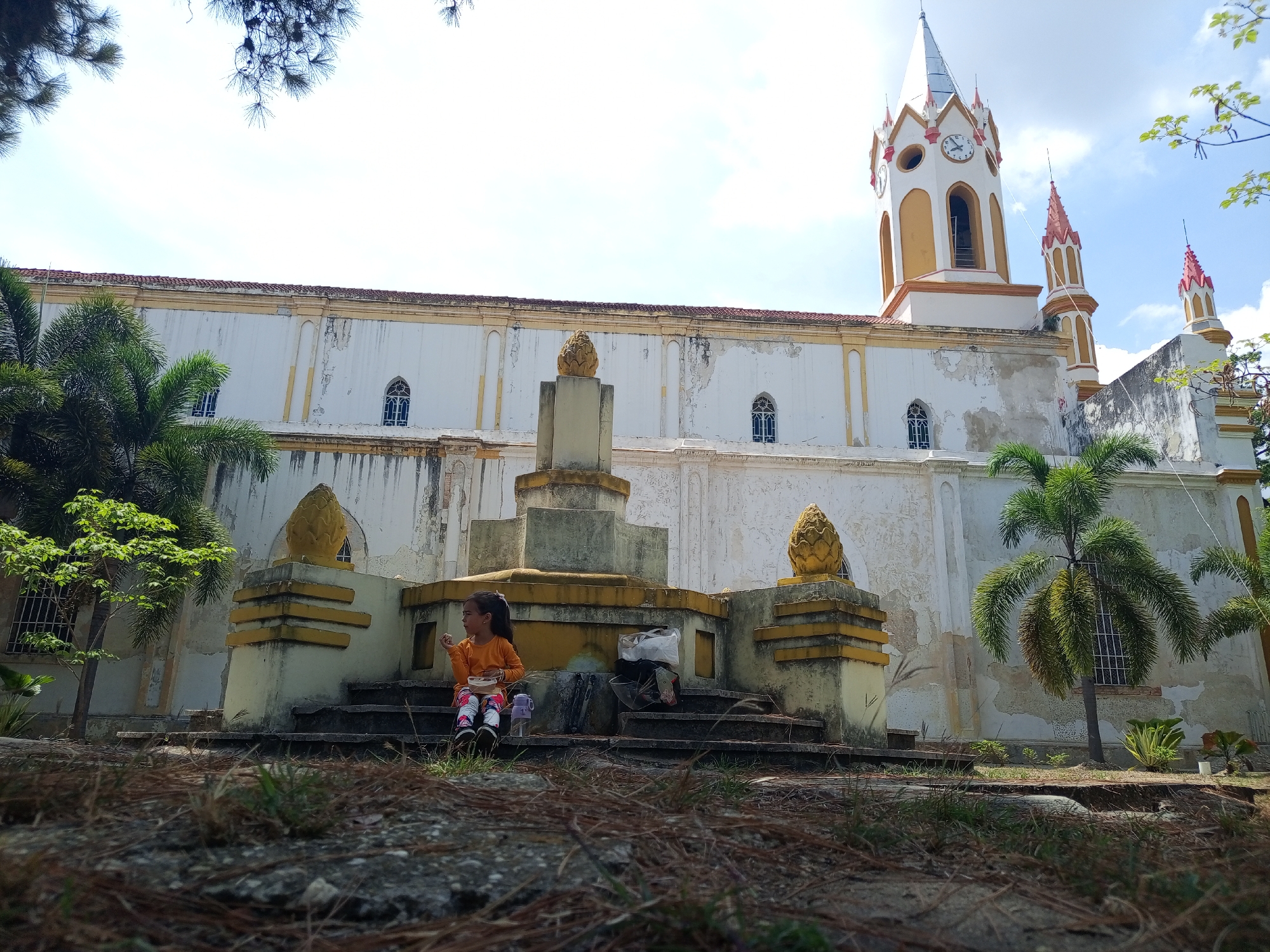 The scene is an eye-level outdoor shot of a church in Tinaquillo, Venezuela, with a girl sitting on the steps leading to a structure in front of the church. The church is a large, aged, white building with a yellow trim and a clock tower with red detailing. The building shows signs of wear and age. In front of the church is a multi-tiered stone structure decorated with pineapple-shaped ornaments.
A young girl with dark hair is sitting on the steps of this structure. She wears a long-sleeved orange shirt and colorful pants. She is holding something in her hand near her mouth, suggesting she is eating. A water bottle and a plastic bag with items are next to her.
The foreground consists of the ground covered with grass and pine needles. The sky is visible at the top of the frame with scattered clouds and some tree branches framing the shot. Palm trees and other green foliage are situated around the church, contributing to the landscape.